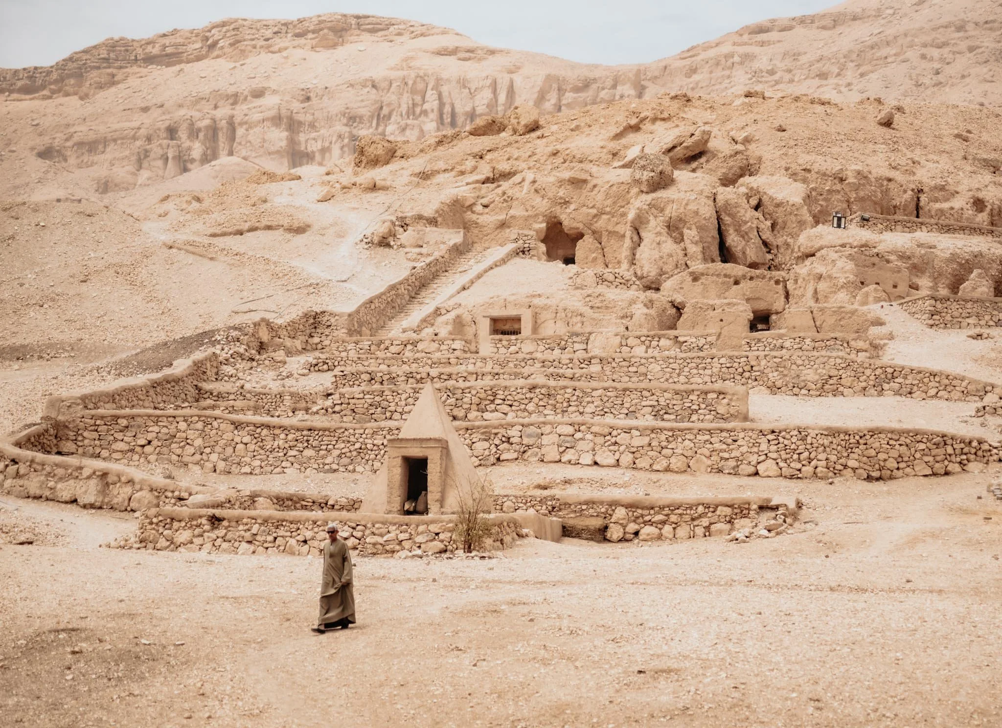 Ancient tombs of the scribes in Luxor, carved into a rocky hillside with terraced stone walls, set against a desert landscape with mountains in the background. A person dressed in traditional Middle Eastern clothing walks in the foreground.