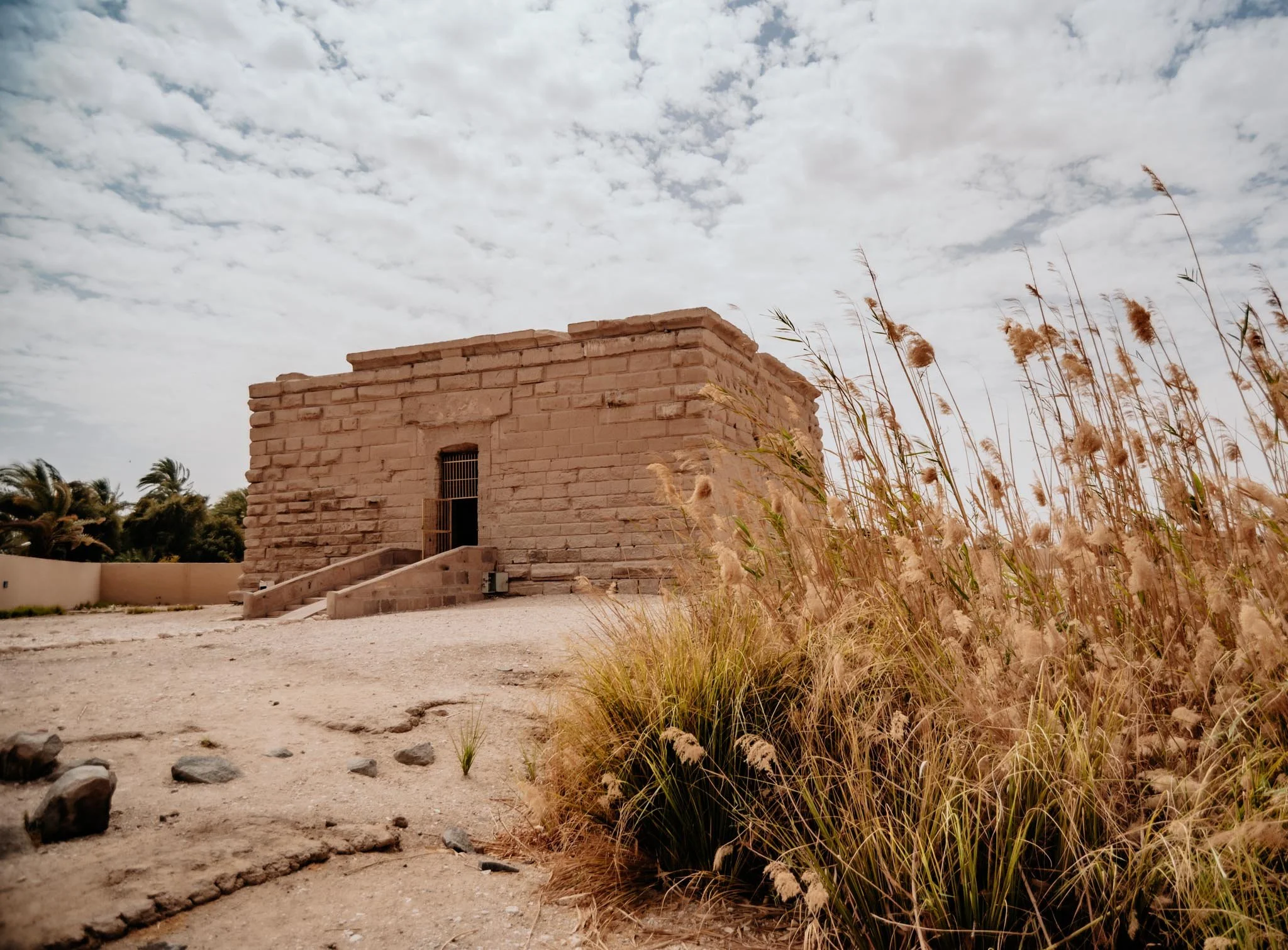 Temple dedicated to Isis in Luxor surrounded by desert landscape and tall grasses under a partly cloudy sky.