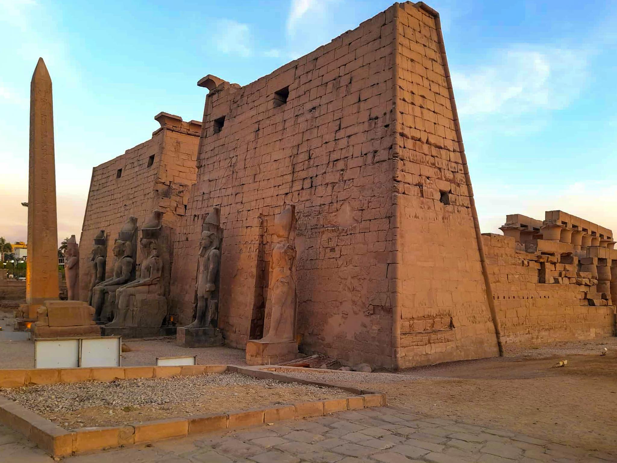 Ancient magical Luxor Temple with large stone walls, seated stone statues, and a tall obelisk under a blue sky.