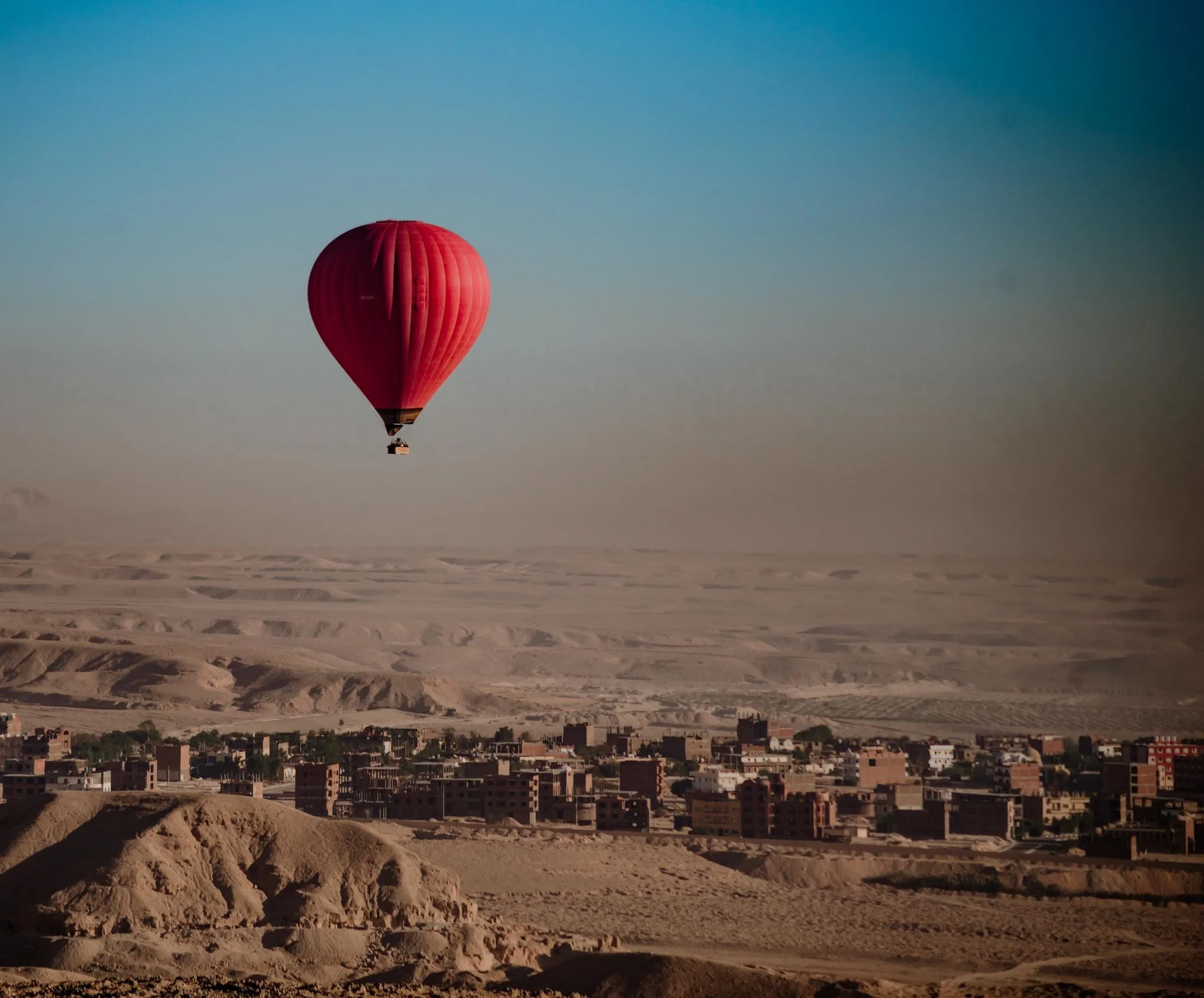 A hot air balloon with a reddish-pink colour floating above a cityscape, set against a sky that transitions from blue to a lighter hue near the horizon early morning in Luxor