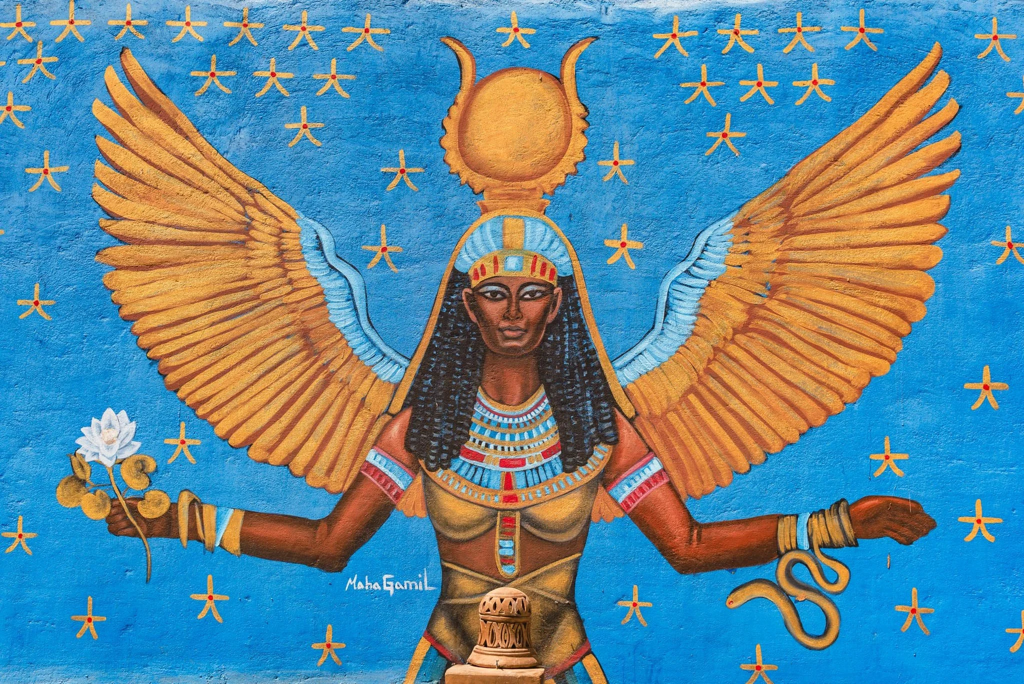 A colorful painting of an Egyptian deity with large golden wings, dark skin, and black curly hair. The figure wears traditional Egyptian jewelry and headgear, with a sun disk and wings above the head. The background is bright blue with small golden m