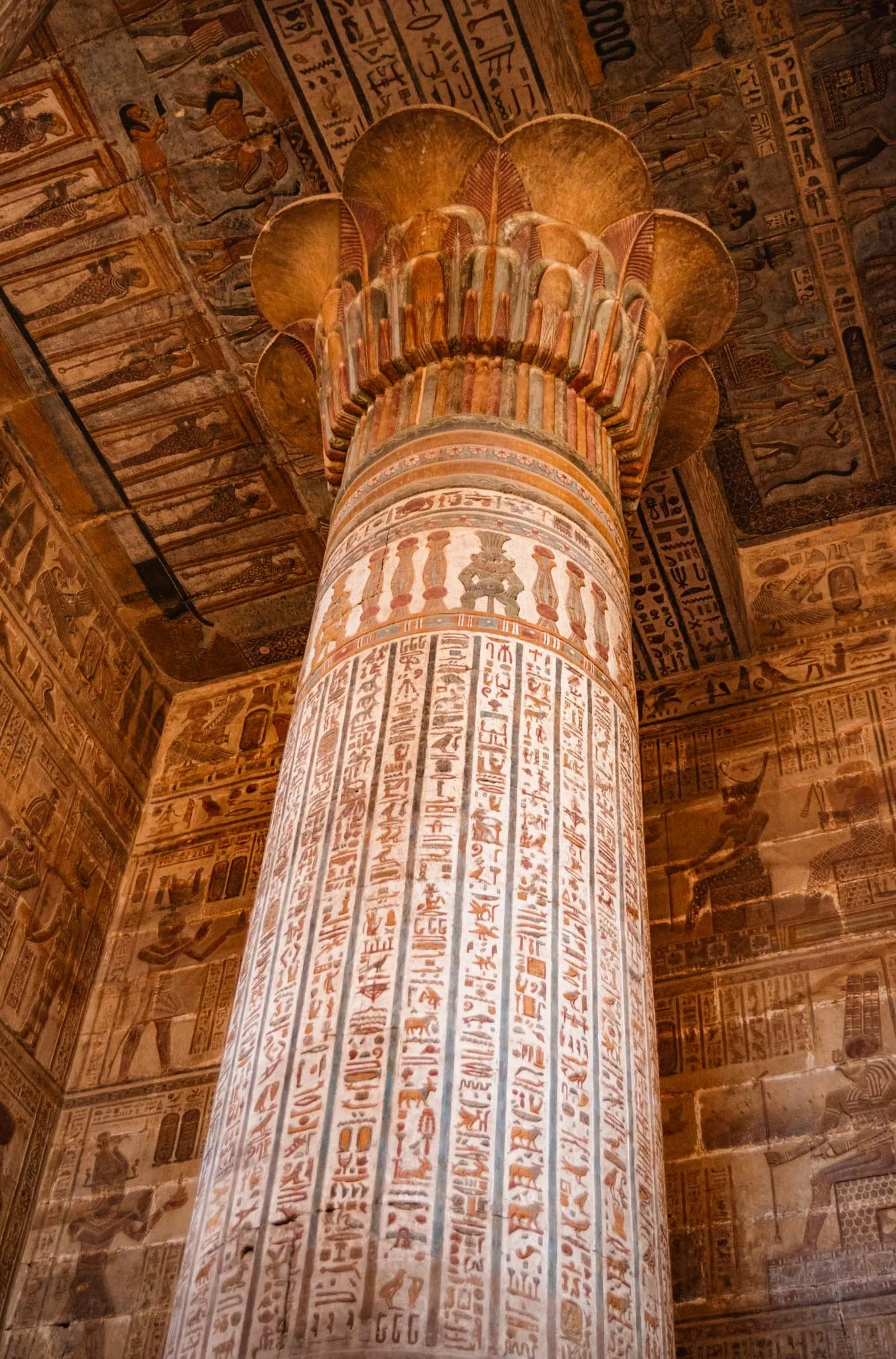 Ancient Egyptian temple interior with a large decorated column covered in hieroglyphics and painted hieroglyphic inscriptions on the walls and ceiling.