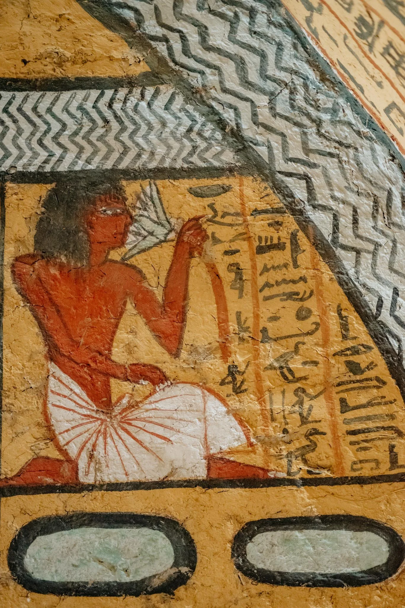 Ancient Egyptian wall painting showing a seated figure with a headdress, holding a staff, with hieroglyphic inscriptions and colorful geometric patterns in the background.