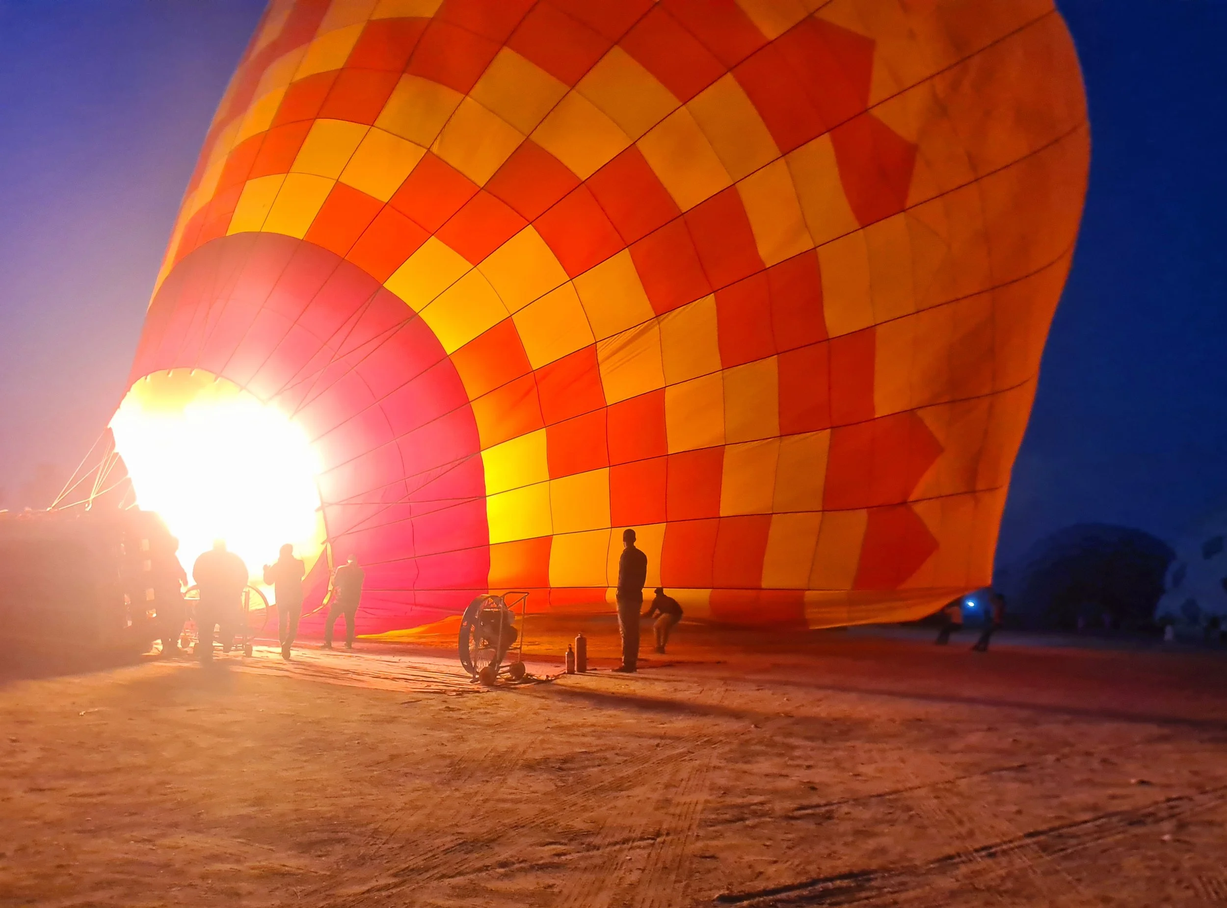 A hot air balloon with a bright orange and yellow checkered pattern on its fabric, illuminated from the flame inside, with silhouetted people gathered around in the early morning in Luxor