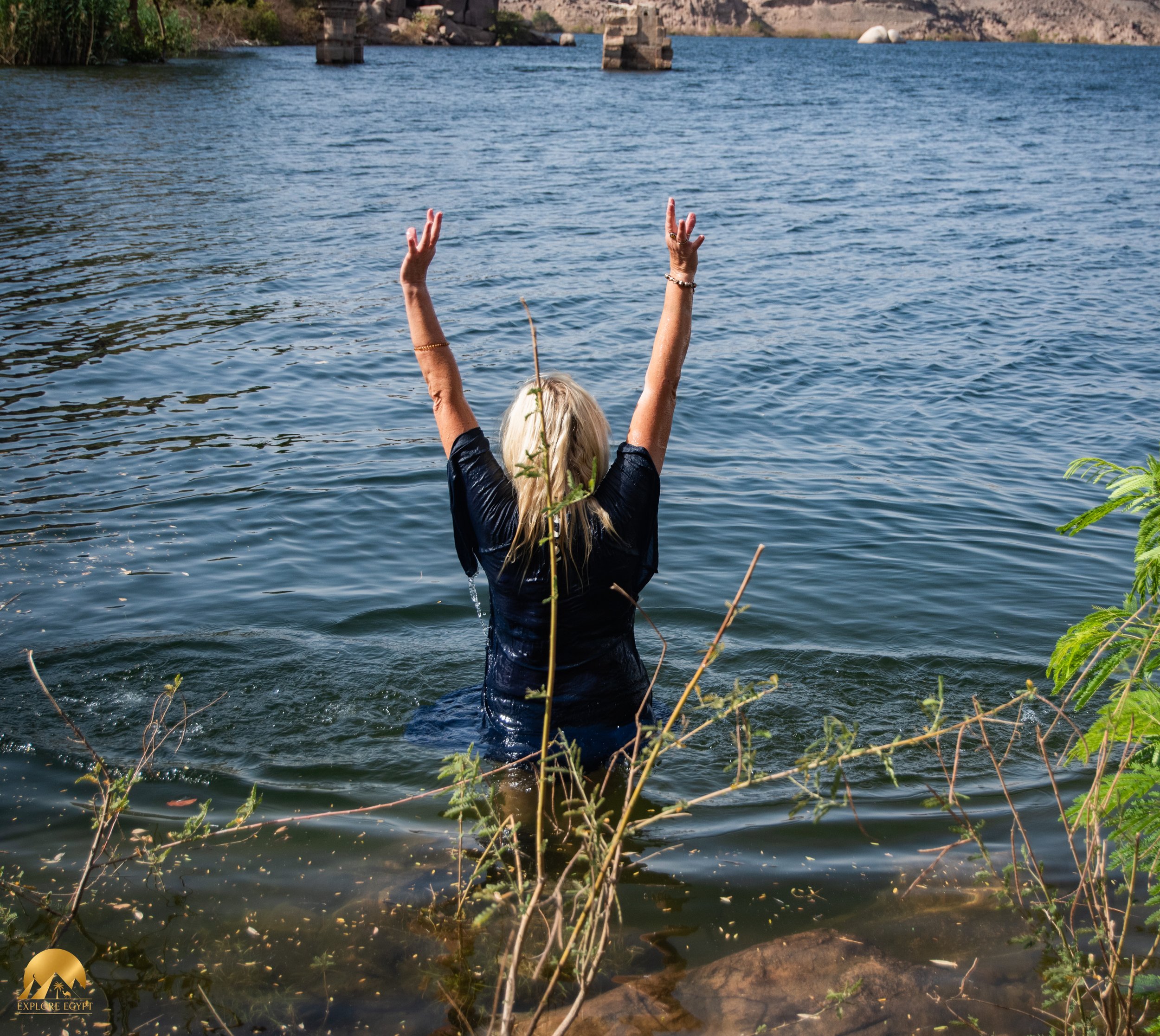 A woman with blonde hair standing in the Nile in Aswan with her hands in the air, facing the calm Nile river with Isis Gate in the background.