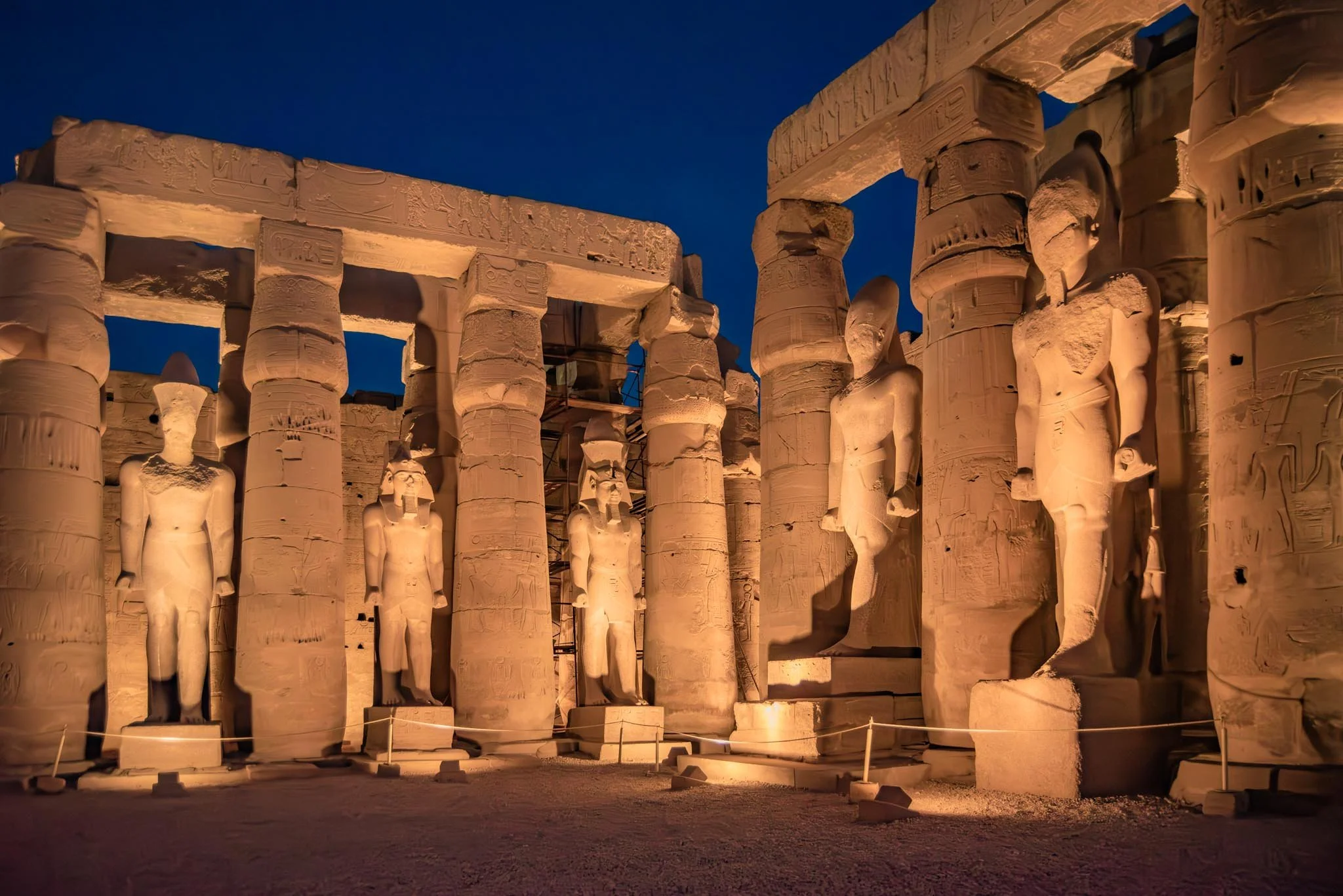 Ancient Egyptian stone ruins illuminated at night in Luxor Temple , featuring carved statues of gods and hieroglyphics on columns and lintels.