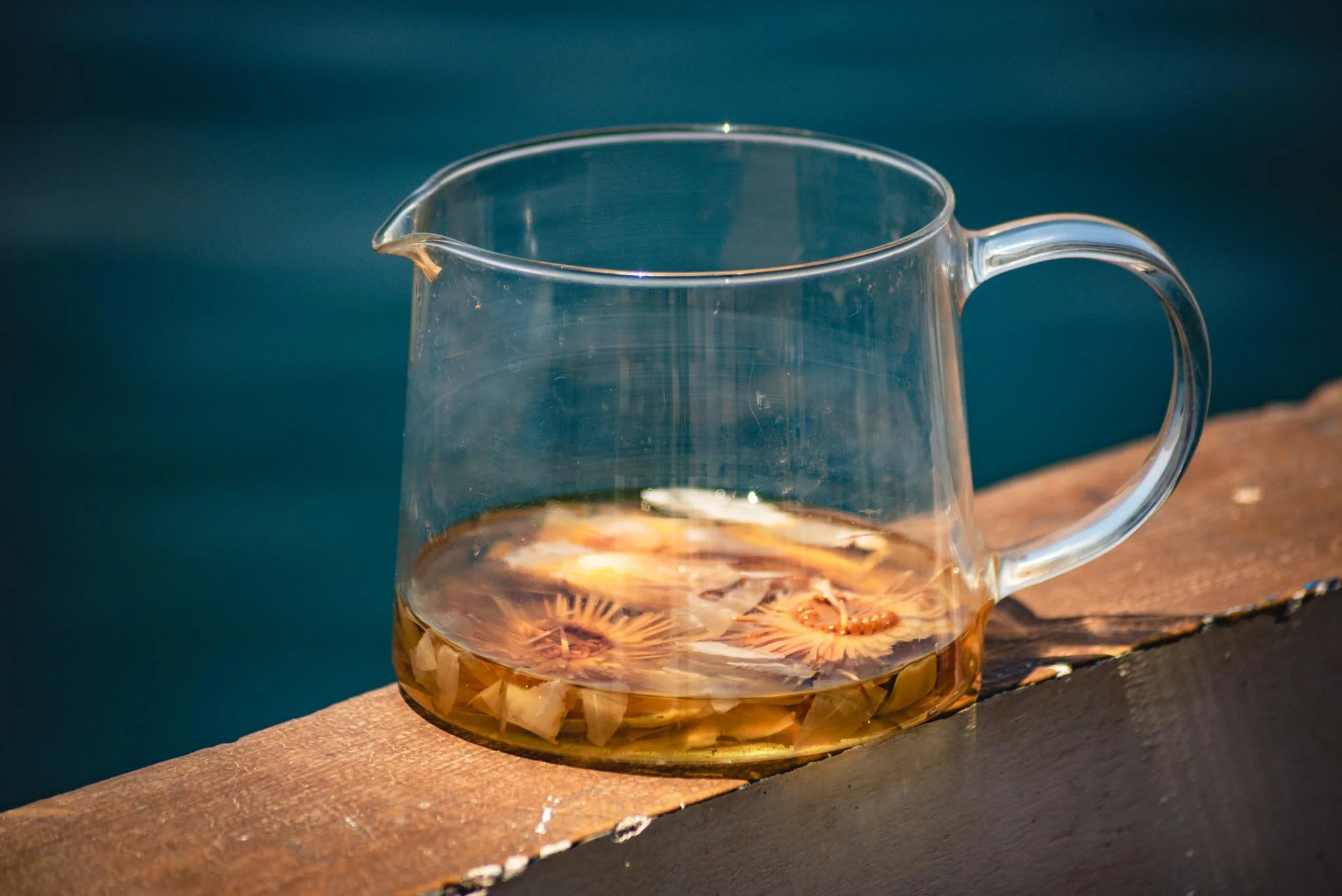 A clear glass pitcher with a small spout and handle, filled with a light amber-colorued liquid and floating blue lotus flowers, placed on a wooden surface with the river Nile in the background