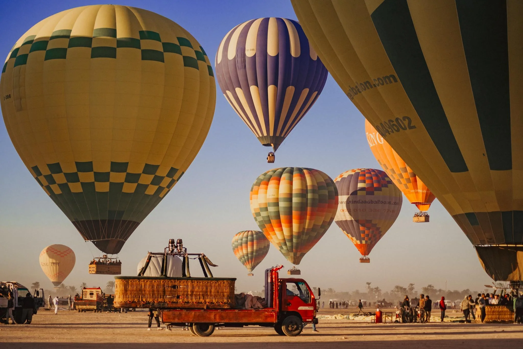 Several hot air balloons in various colours and patterns are floating in the sky during sunrise in Luxor. There are some people on the ground and vehicles, including a red truck, preparing for the hot air balloon ascending