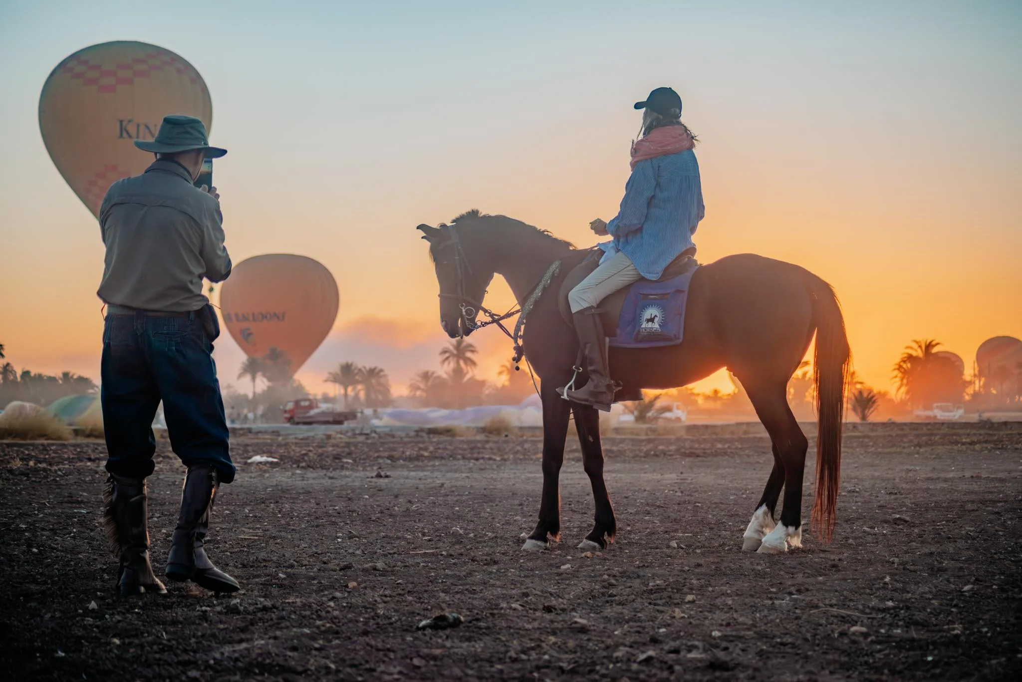 Wonderfully magical scene of a woman on a horse at sunrise in Luxor, with hot air balloons in the sky and a man taking her photo.