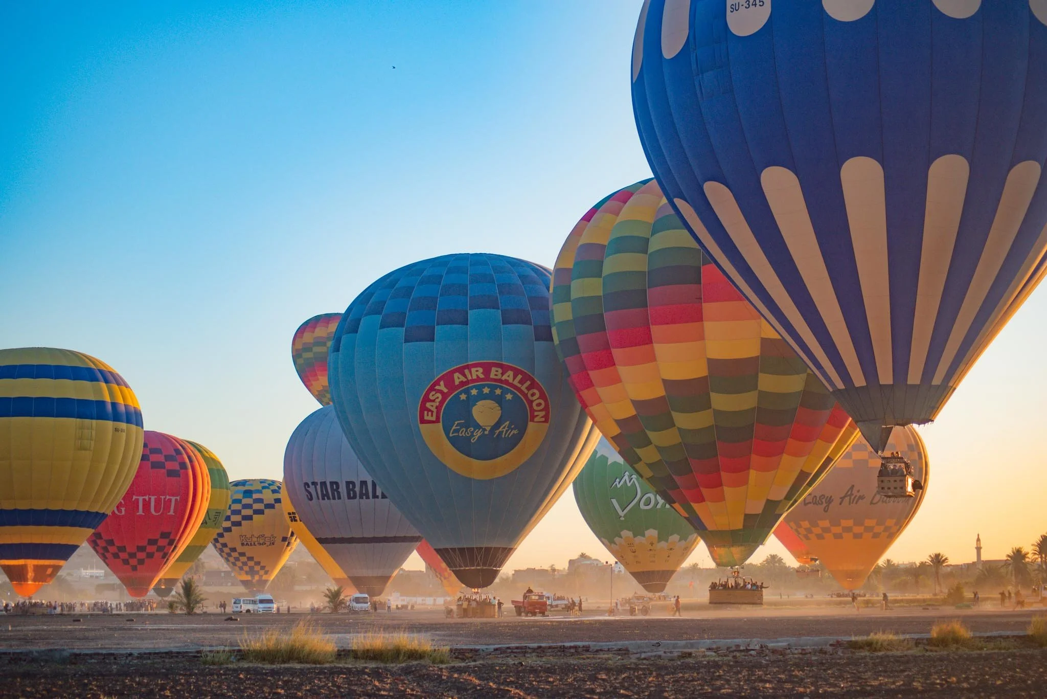 Multiple colourful hot air balloons floating in the sky during sunrise in Luxor, with  a crowd of people on the ground watching the magical display.