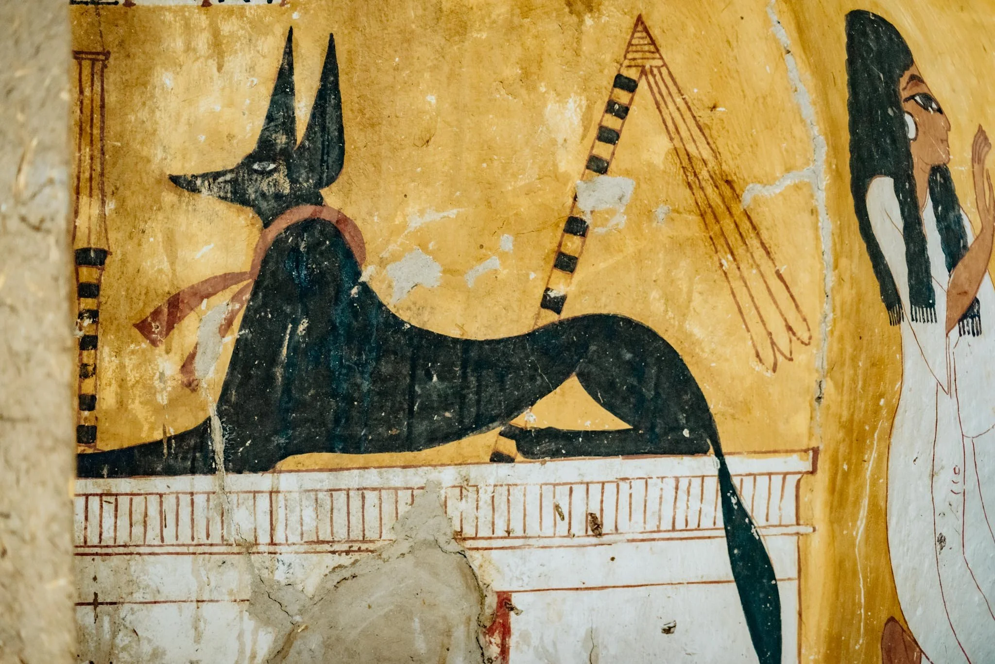 Ancient Egyptian painted illustration of a deity or mythical figure with a falcon head, black headdress, and colorful garments, holding a staff, on a yellow background with hieroglyphic borders.