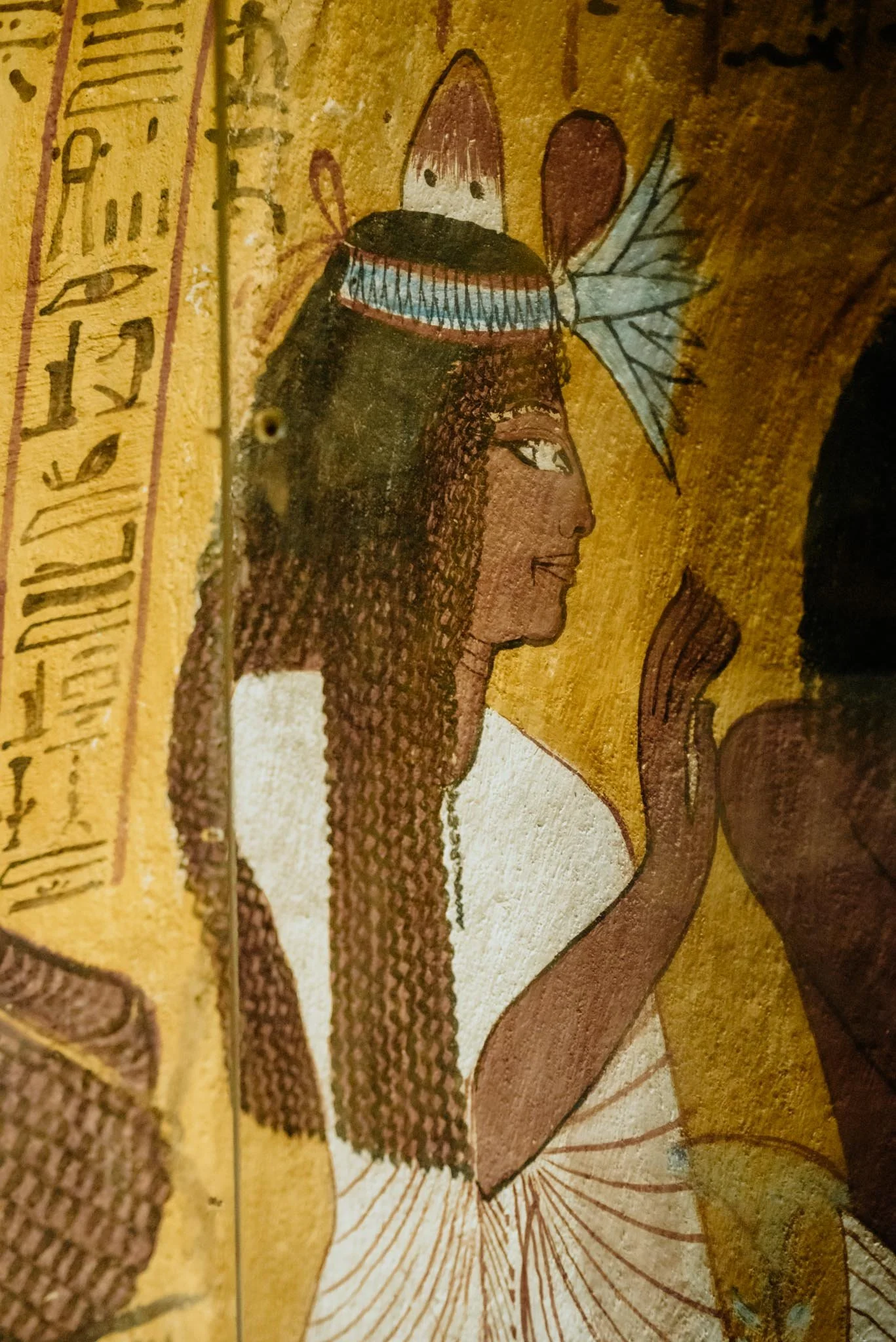 Ancient Egyptian artwork of a woman with dark, curly hair wearing a white garment, with elaborate jewelry and a headdress featuring a reddish-brown element and a blue band, on a yellow background with hieroglyphics.