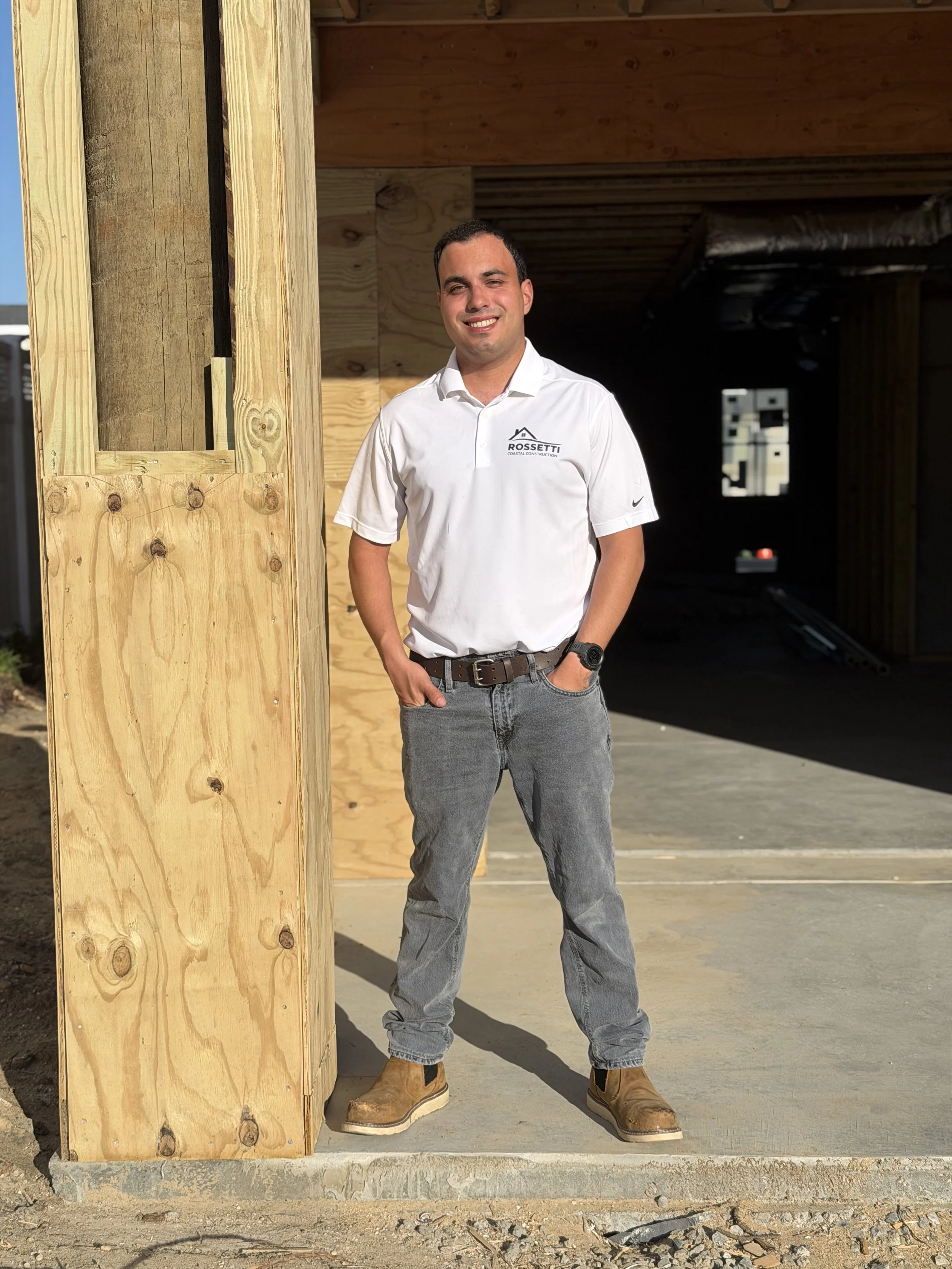 Man standing outside near a wooden column at a construction site, wearing a white polo shirt with a company logo, gray jeans, and tan work boots.