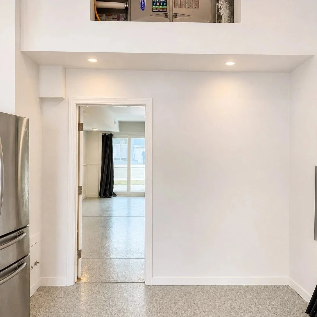 View of a bright room with sliding glass door and black curtains, seen through an open doorway from a kitchen area with a stainless steel refrigerator.