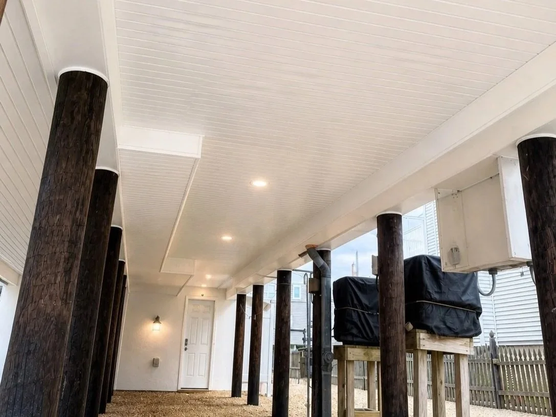 Under a house with a white ceiling and wall, wooden support posts, and a gravel ground, two items covered with black tarps are on a wooden platform, with a white door and exterior siding visible in the background.
