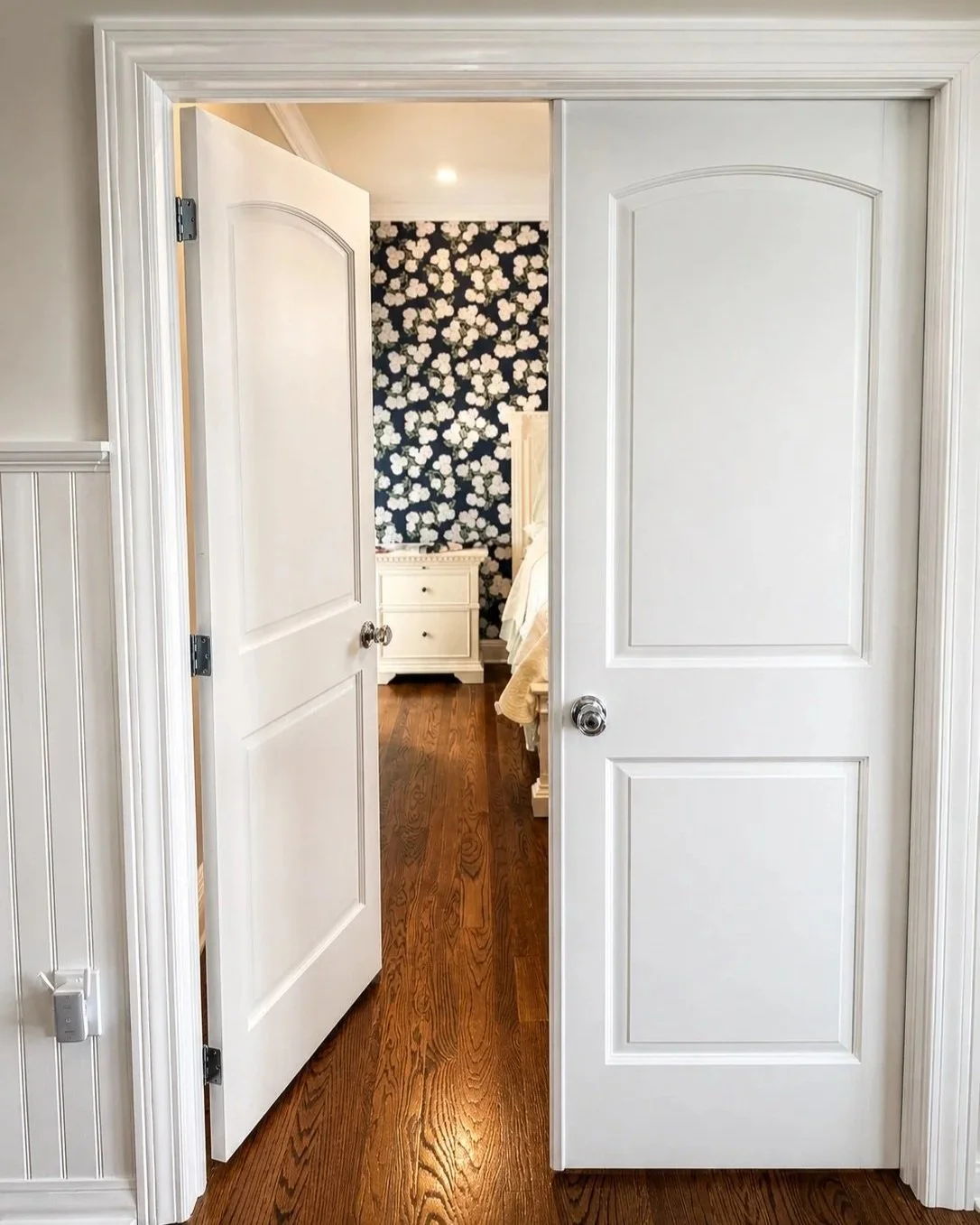 Double white doors partially open revealing a bedroom with wooden flooring, floral wallpaper, a white nightstand, and a bed.