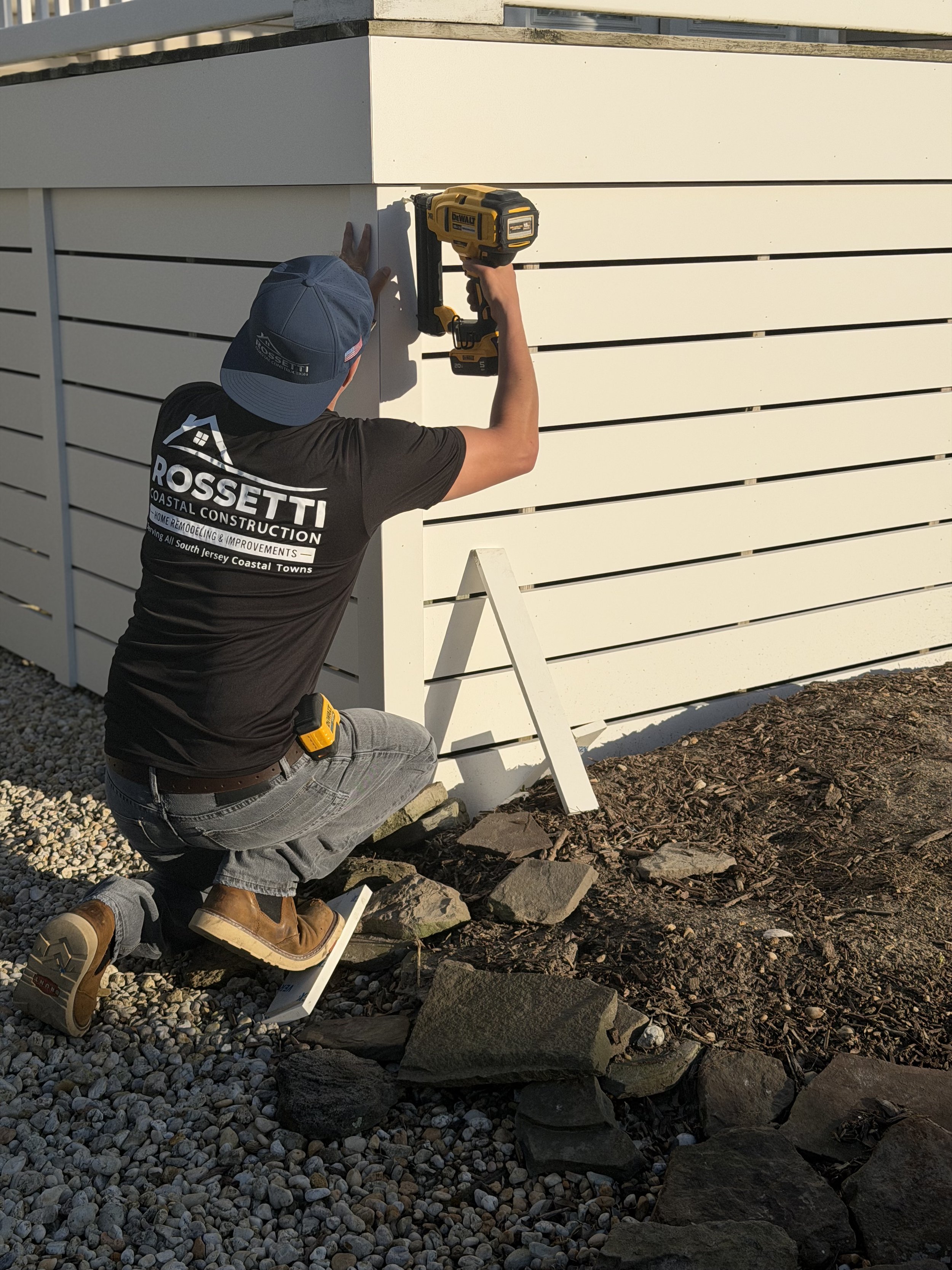 Person kneeling on gravel ground, using a cordless drill on a white horizontal wooden slat wall. The person is wearing a blue cap, a black T-shirt with white and red text, gray jeans, and brown boots. The wall is part of an outdoor structure, and there is a white step ladder nearby.