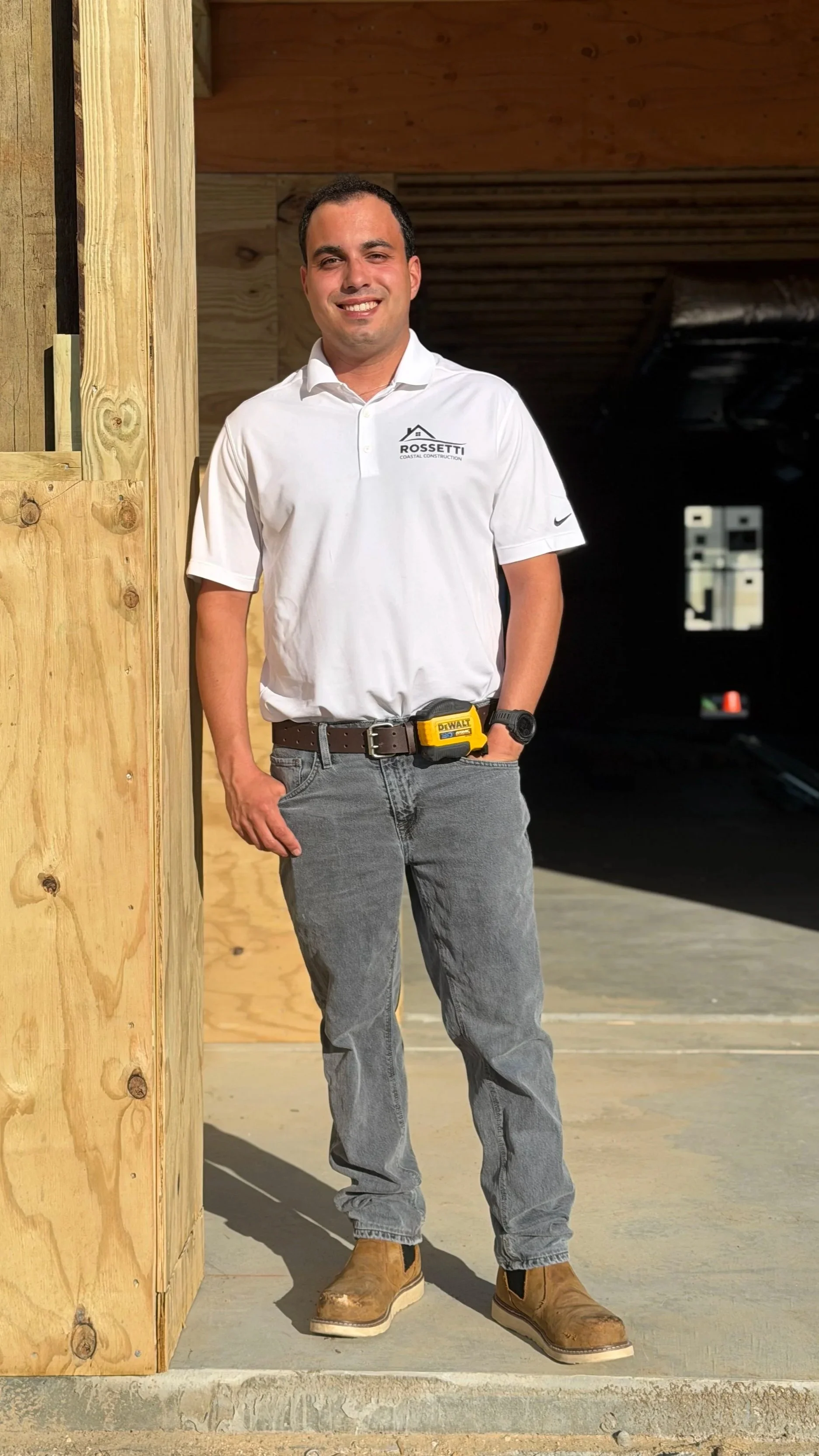 A man standing inside a construction site, wearing a white polo shirt with a company logo, gray jeans, work boots, and a yellow belt tool. The background shows wooden framing and partially completed walls.