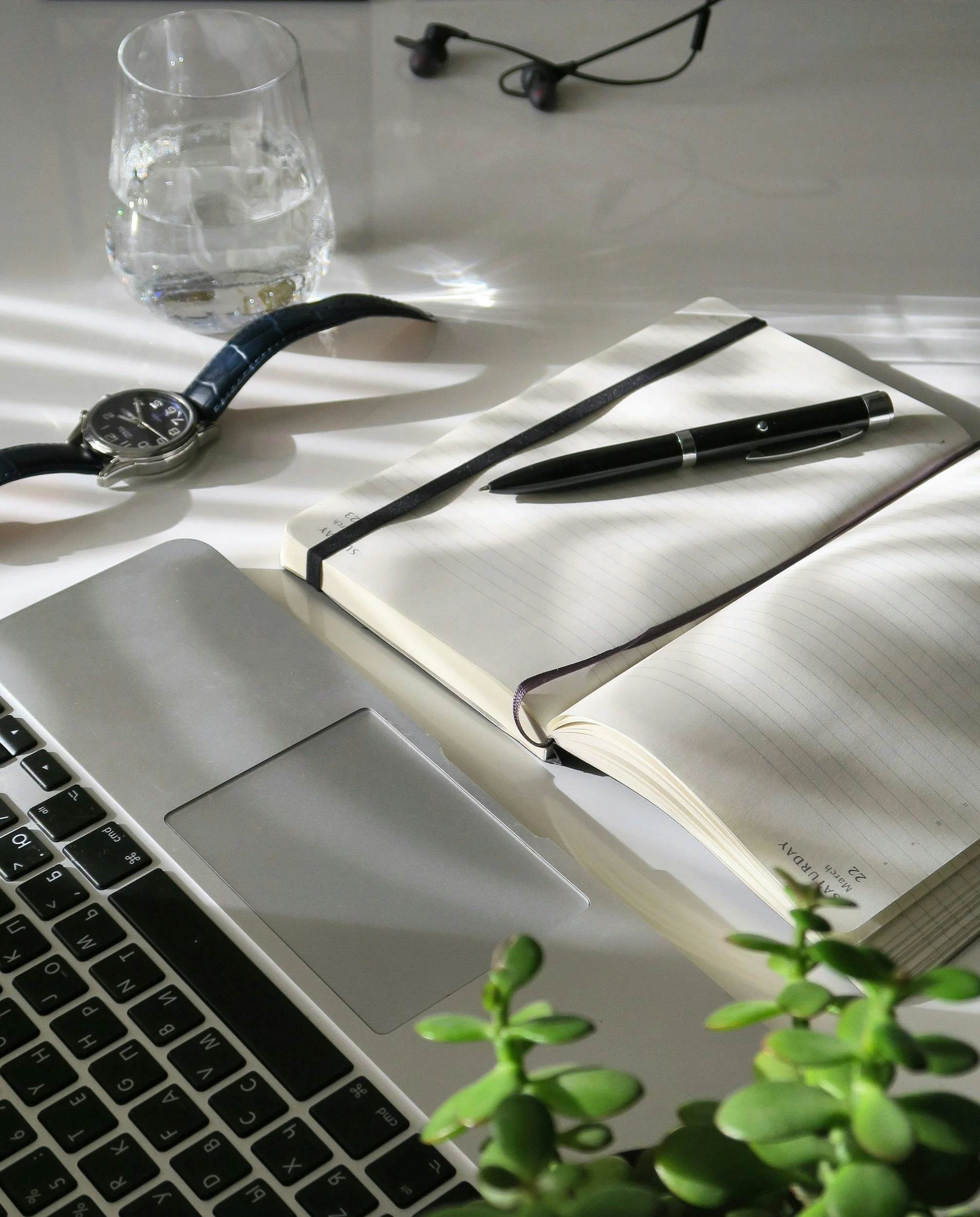 A workspace with a laptop, an open notebook with a pen on top, a wristwatch, a glass of water, a pair of in-ear headphones, and some green plant leaves.
