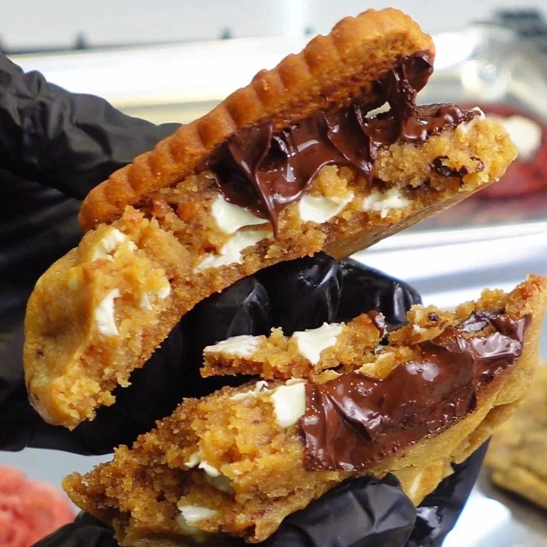 Close-up of a checkerboard cookie sandwich dessert with peanut butter filling, topped with chocolate spread and crushed cookies, held by a hand in a black glove.
