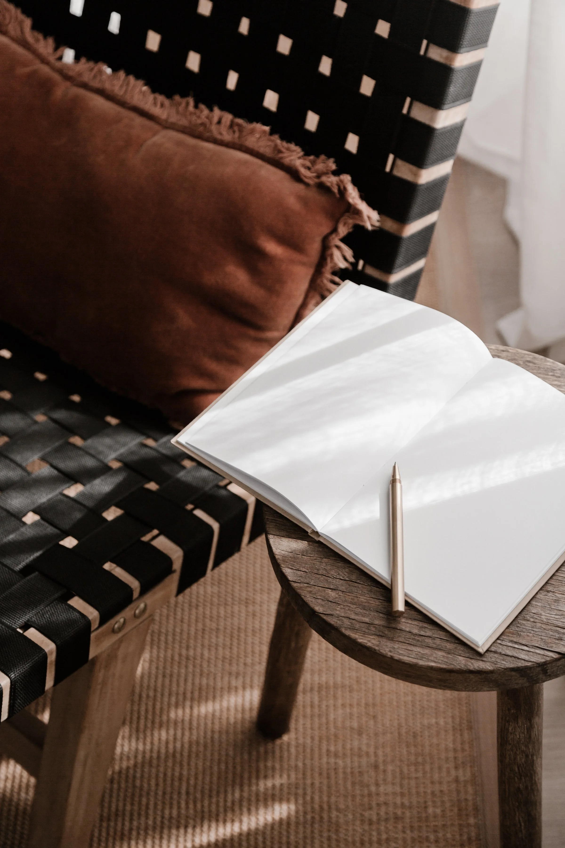 An open blank notebook with a pen on a wooden side table next to a chair with a brown cushion and patterned black and beige armrest, with sunlight coming through a window