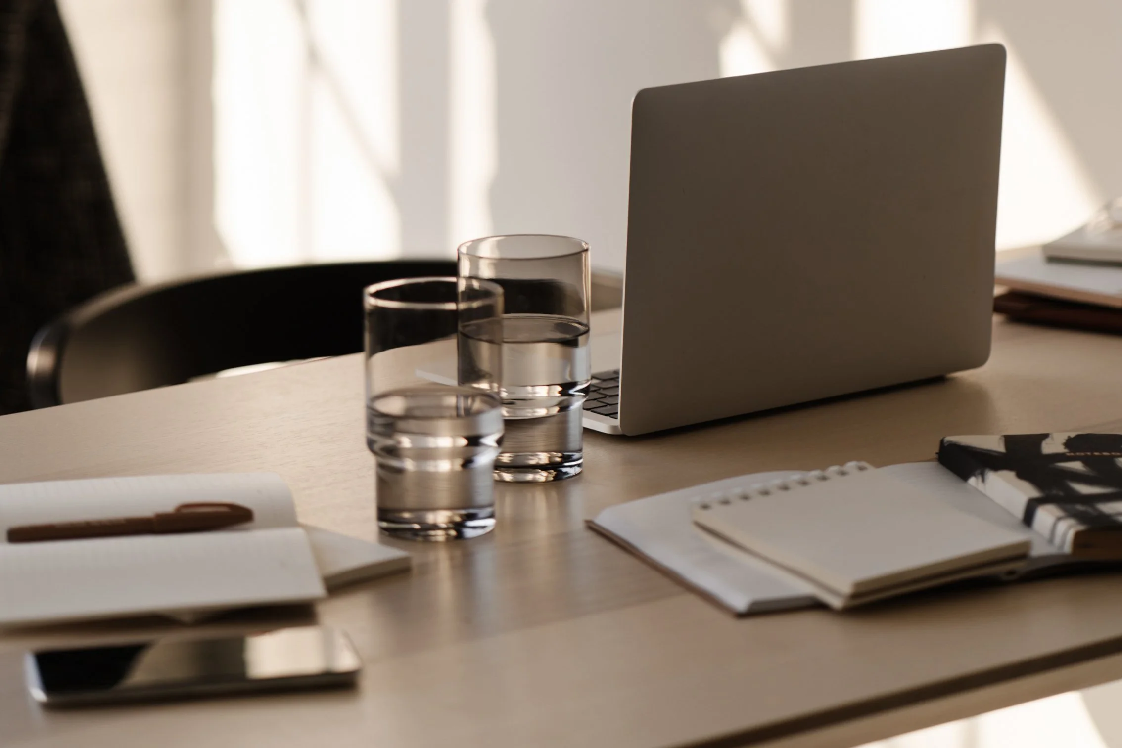 A cluttered wooden table with a closed laptop, two glasses of water, notebooks, a pen, and a smartphone in a well-lit room.