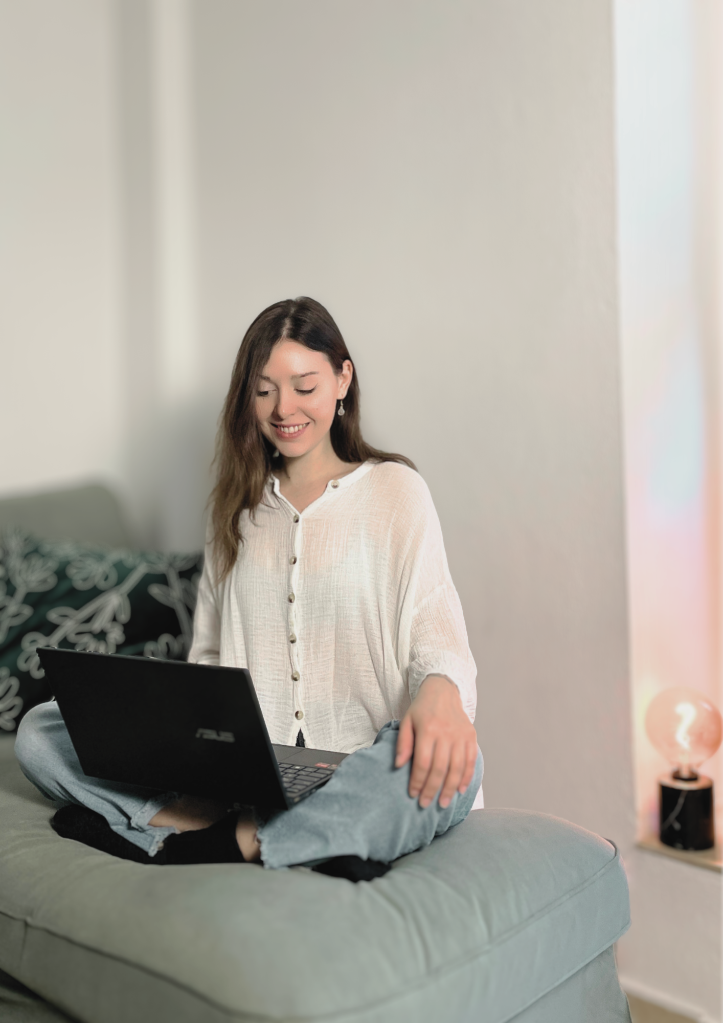 Junge Frau sitzt entspannt auf einer Couch, arbeitet an einem Laptop und lächelt in einem hellen, modernen Raum.