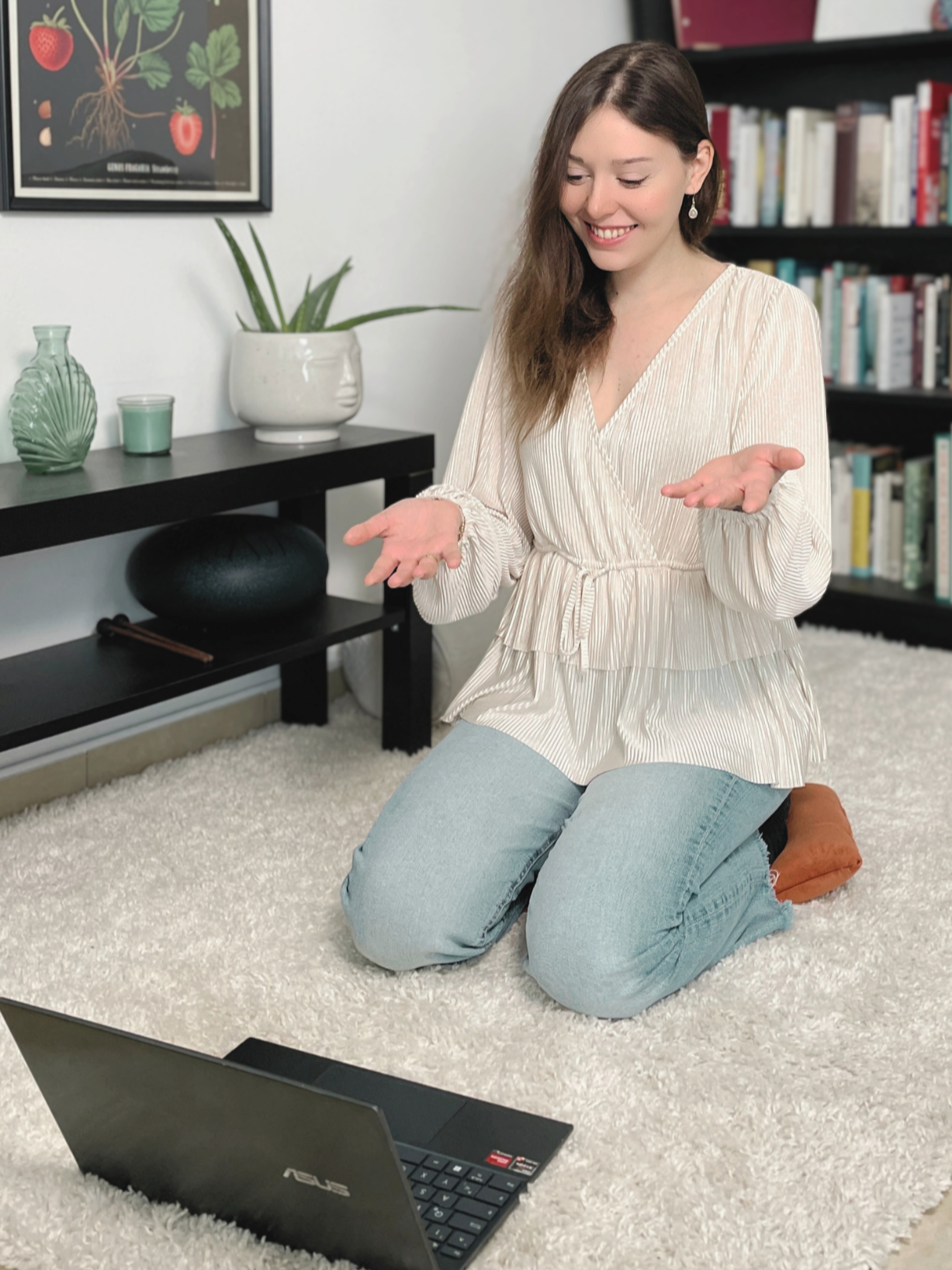 Eine lachende Frau sitzt kniend auf einem weißen Teppich vor einem Laptop. Sie trägt eine beige, gestreifte Bluse und blaue Jeans. Im Hintergrund sind Bücherregale, eine Pflanze in einer weißen Vase, und Dekorationsgegenstände auf einem schwarzen Tisch