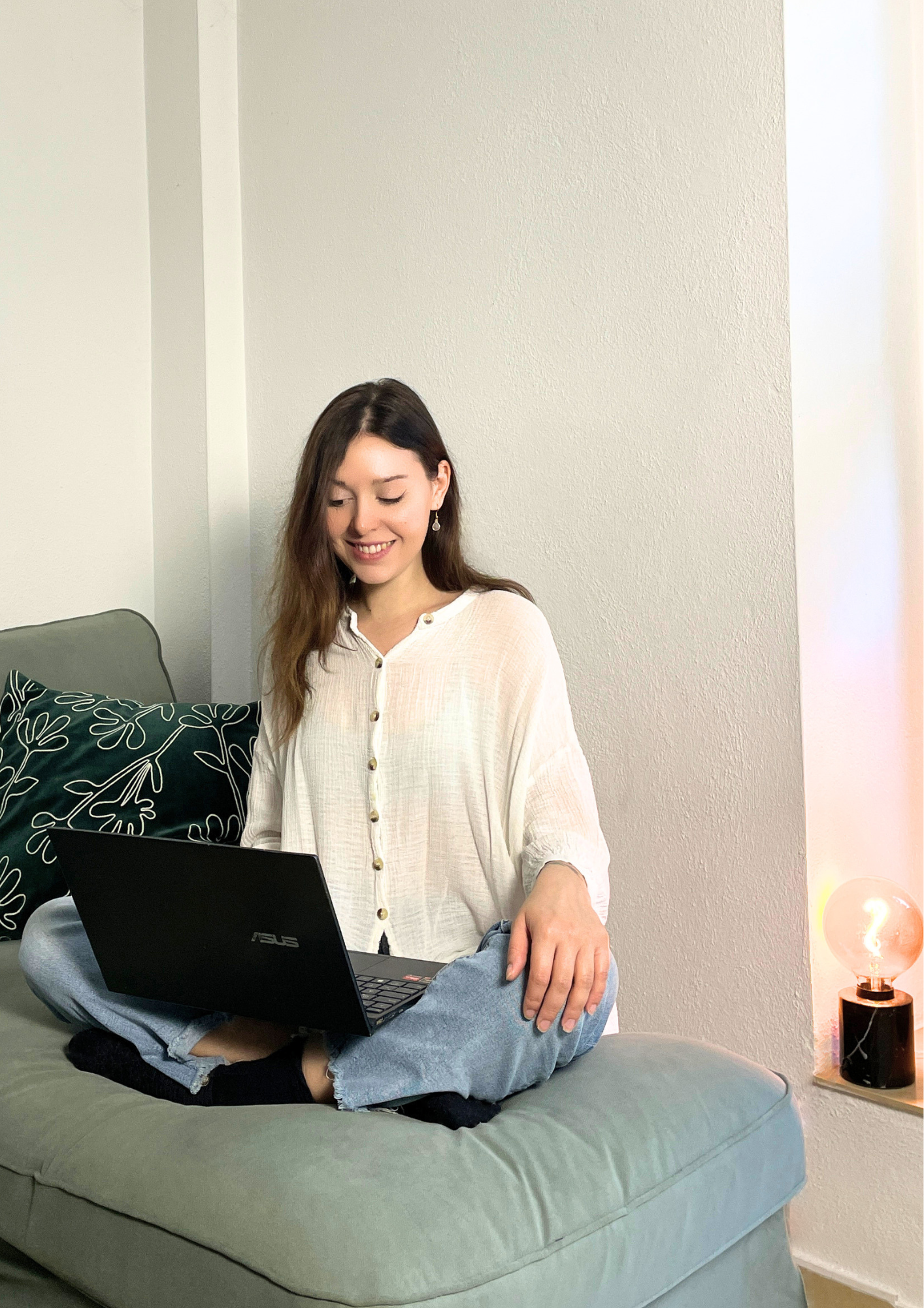 Junge Frau sitzt auf einem Sofa, hat ein Laptop auf den Beinen und schaut darin, lächelt und trägt eine weiße Bluse und Jeans. Im Hintergrund eine Stehlampe mit Glas und Glühbirne sowie ein graues Kissen mit weißen Linien auf einem Sofa.