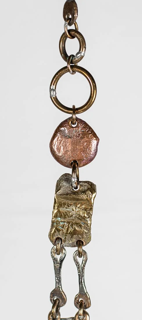 The copper disc is the softest metal in the piece, hammered thin and left to hold its own dull rose against the polished bronze above it.