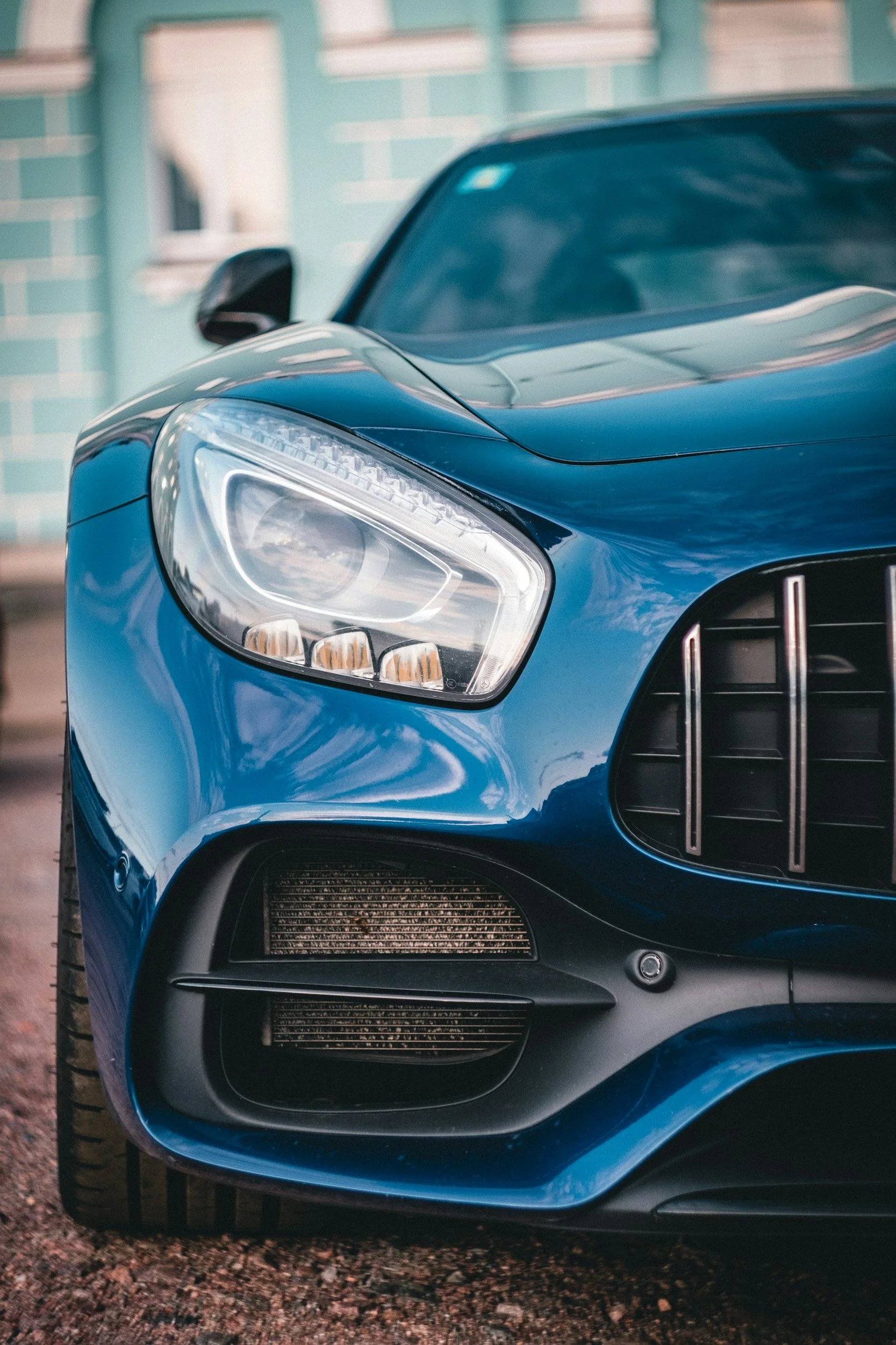 Close-up of the front left side of a shiny blue car, showing the headlight, part of the grille, and wheel, with a green building in the background.