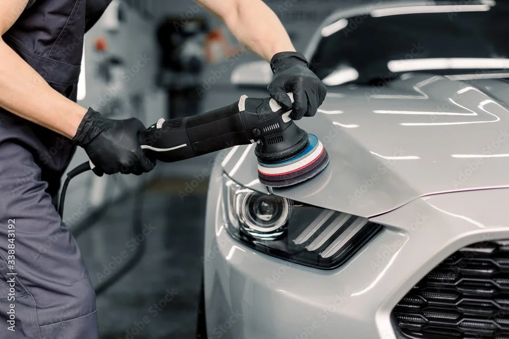 A person using a polishing machine on a silver car's hood in an auto repair shop.