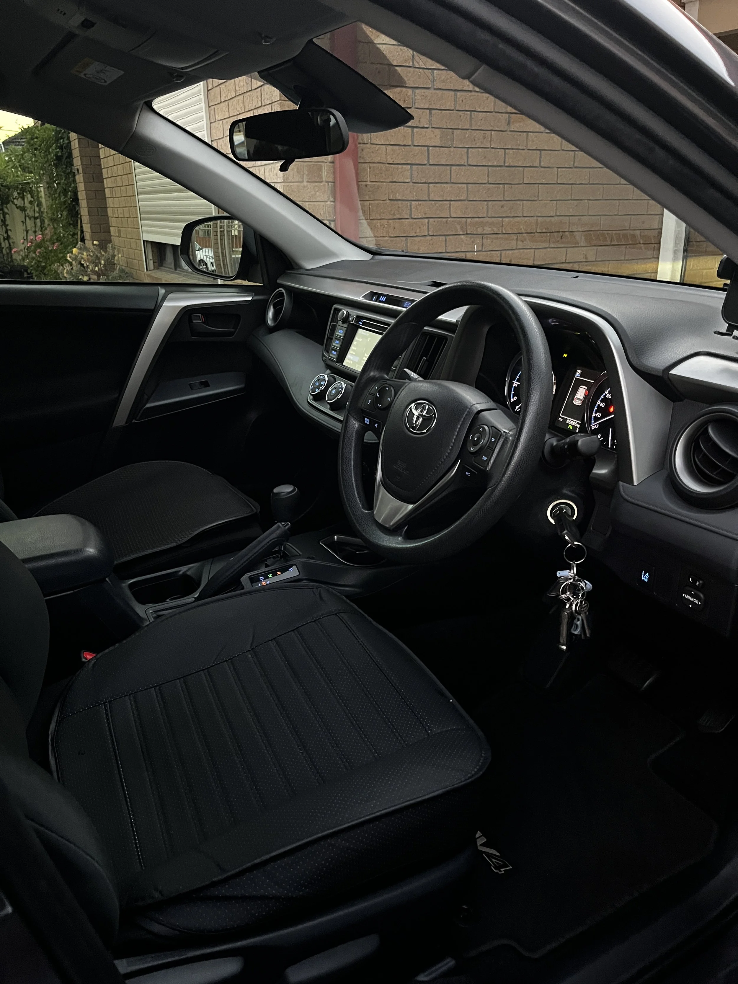 Interior view of a black Toyota vehicle with a dashboard, steering wheel, gear shift, keys in the ignition, and a black seat cover, parked in front of a brick wall.