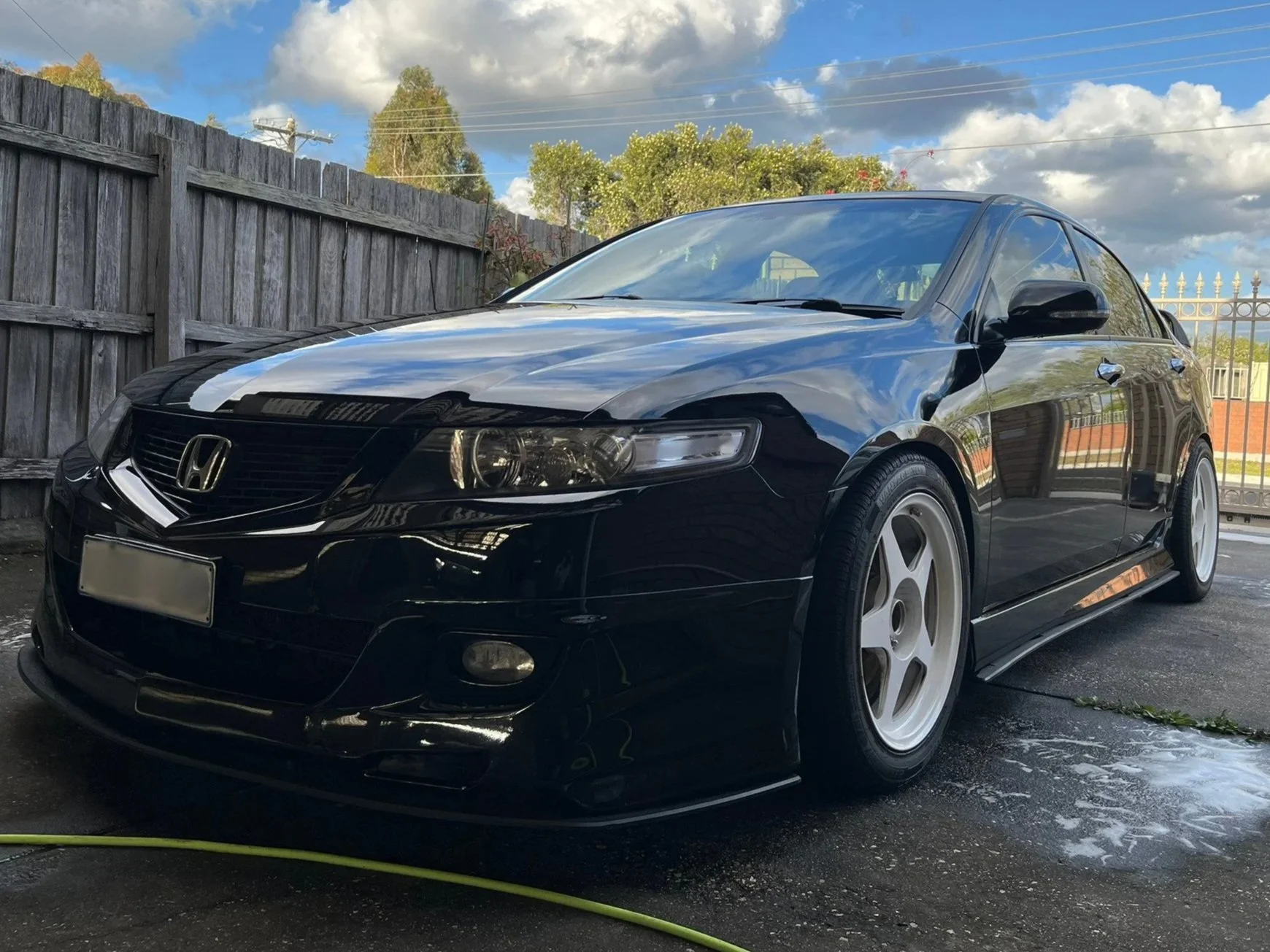 Black Honda car parked outdoors with a wooden fence, cloudy sky, and trees in the background, showing a white racing stripe on the hood.
