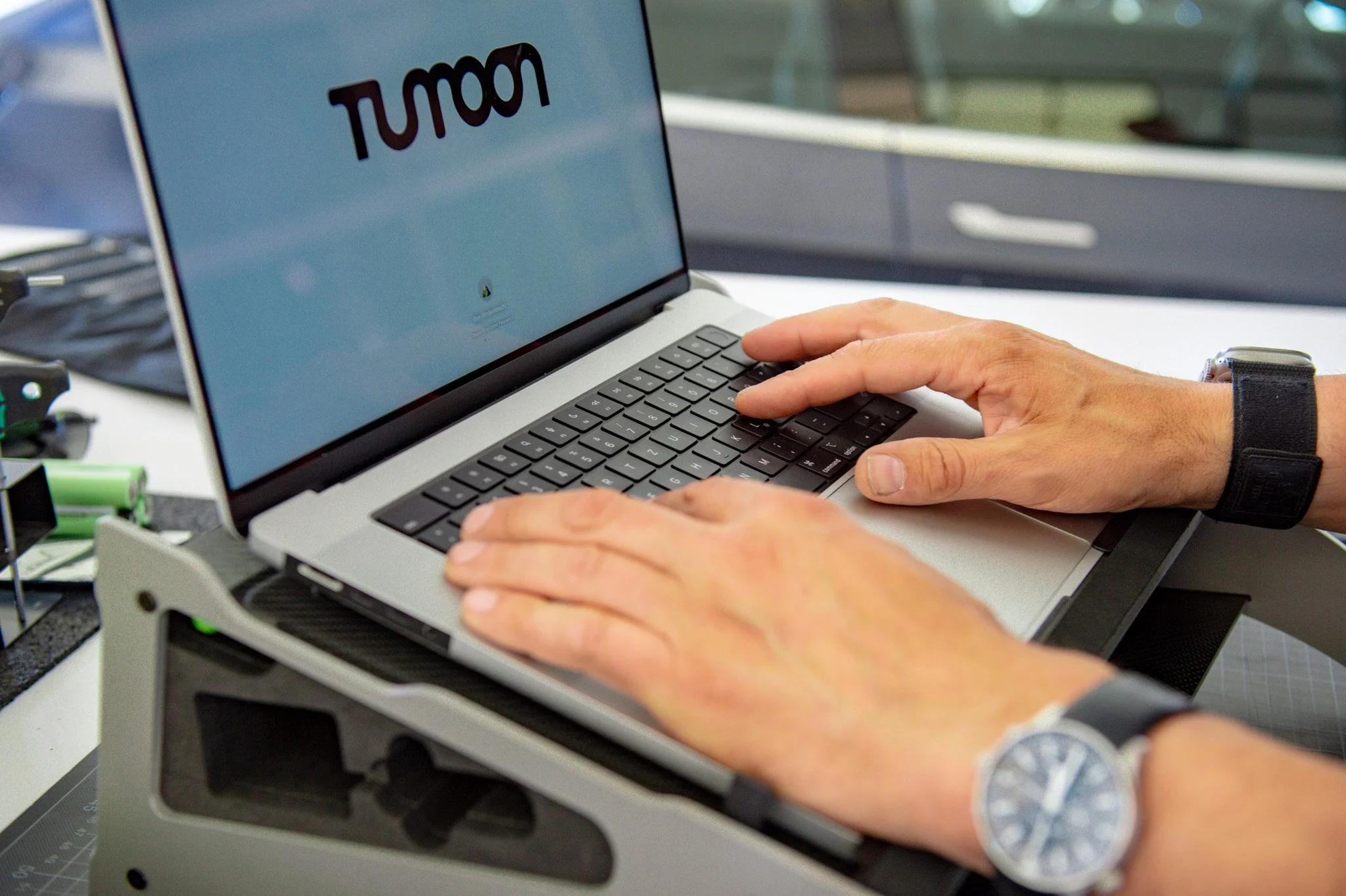 Nahaufnahme einer Person, die an einem Laptop arbeitet, mit sichtbarer Uhr am Handgelenk, auf einem Schreibtisch, mit dem Bildschirm, auf dem das Wort 'TUMON' zu sehen ist.