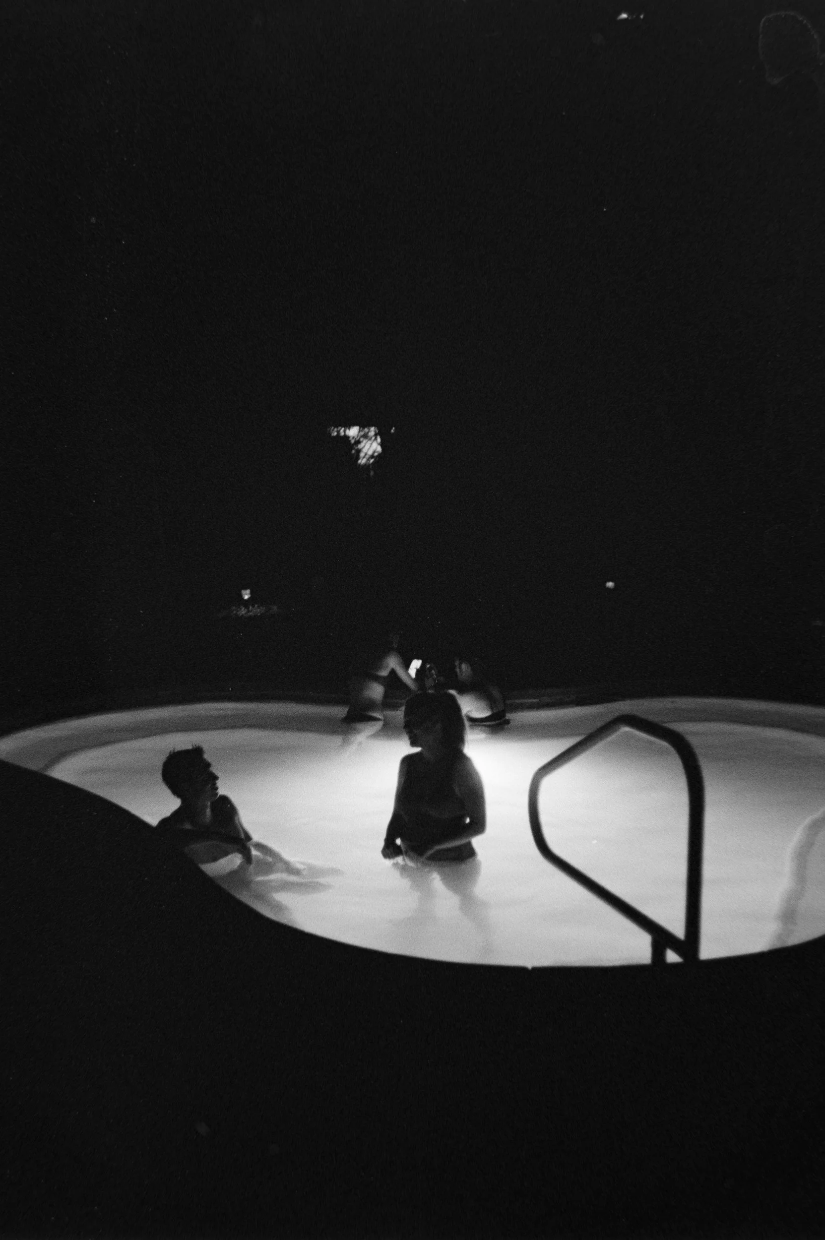 Children standing in a shallow pool at night, illuminated by underwater lights, with dark sky and trees in the background.