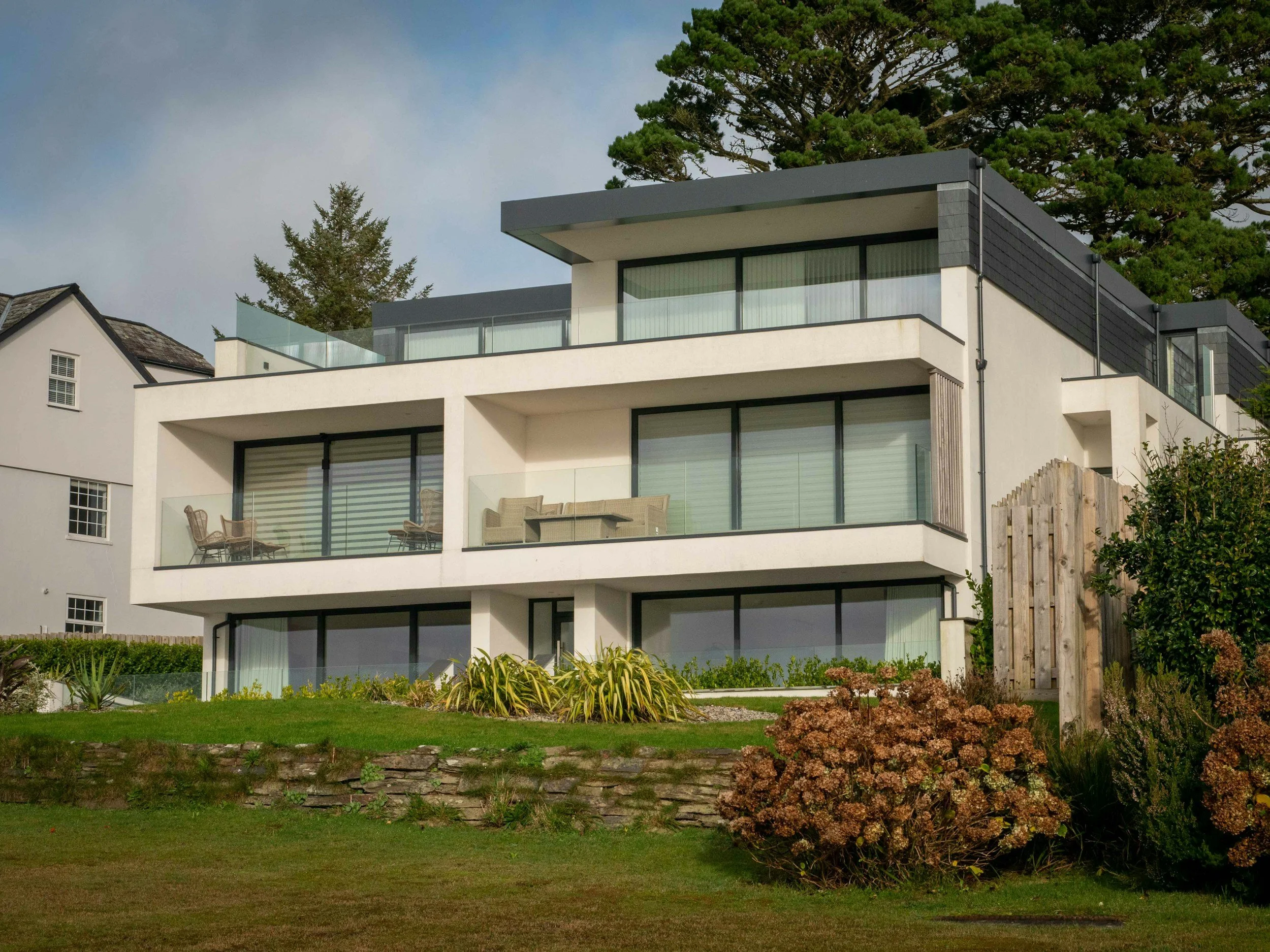 Modern white multi-story house with large glass sliding doors, balconies, and a landscaped garden with lush green grass and various plants and bushes.