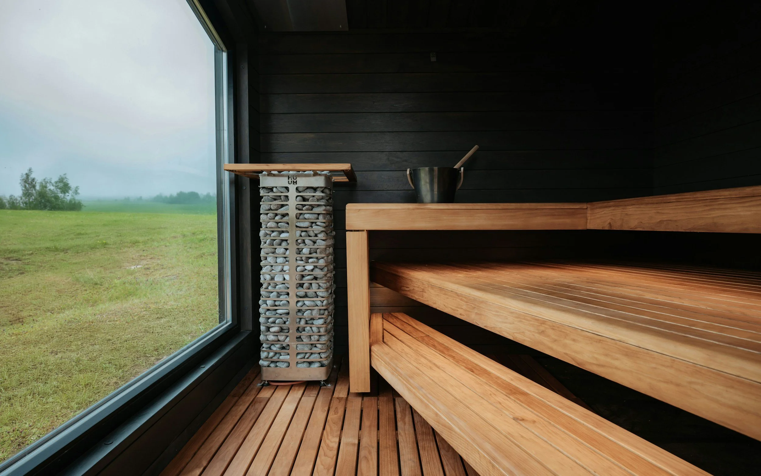 Interior of a sauna with wooden benches, a black exterior wall, and a large window showing an outdoor grassy landscape and overcast sky.
