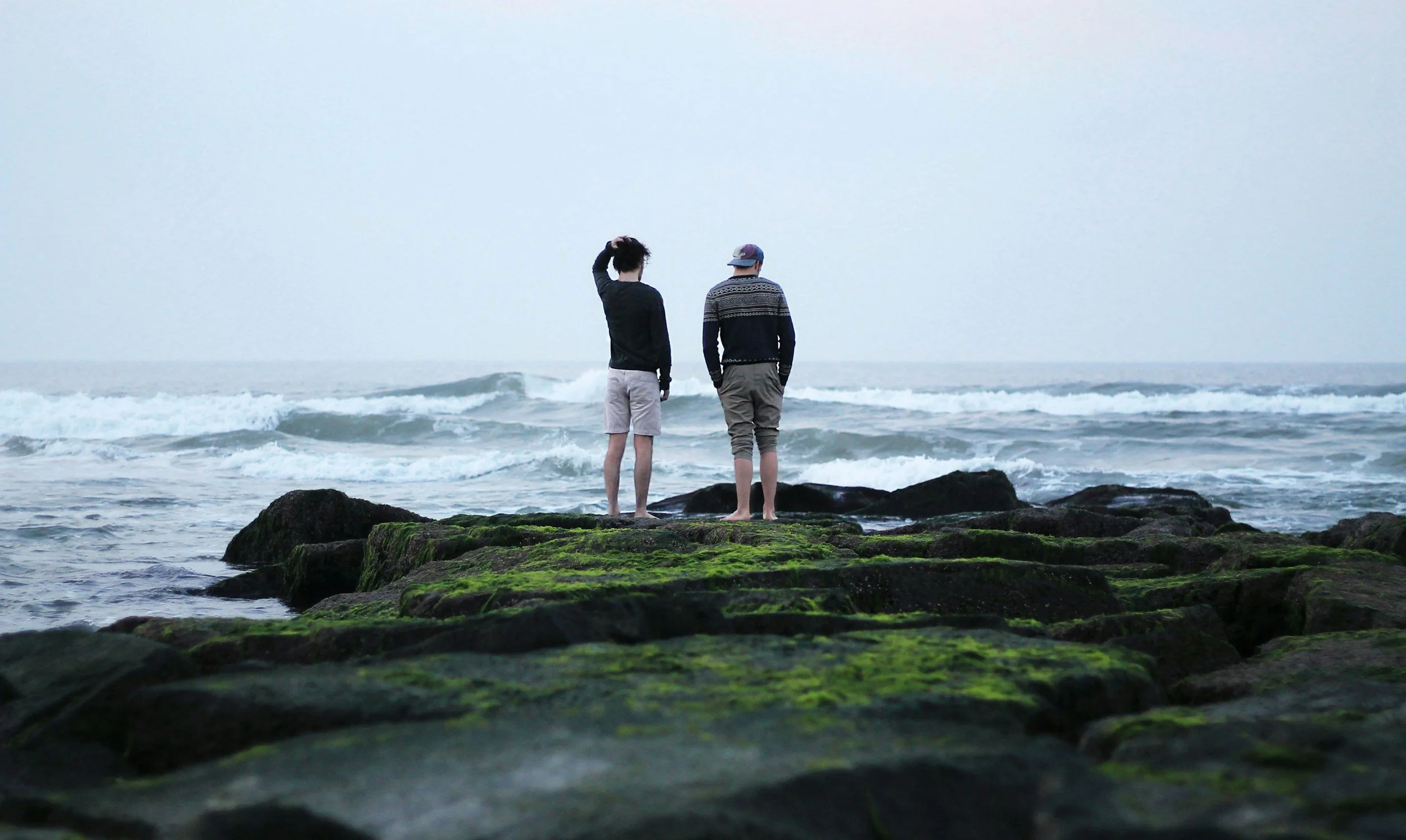 Two people standing on moss-covered rocks near the shoreline, looking at the ocean with small waves, during overcast weather.