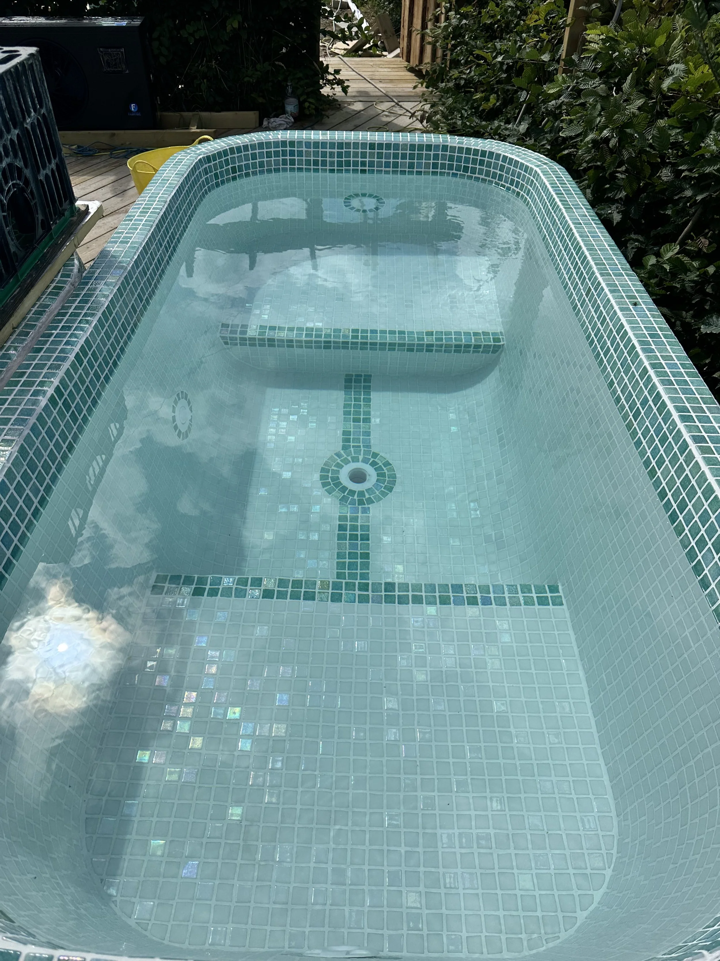 A small above-ground hot tub with teal mosaic tile interior, filled with water, situated on a wooden deck surrounded by greenery.