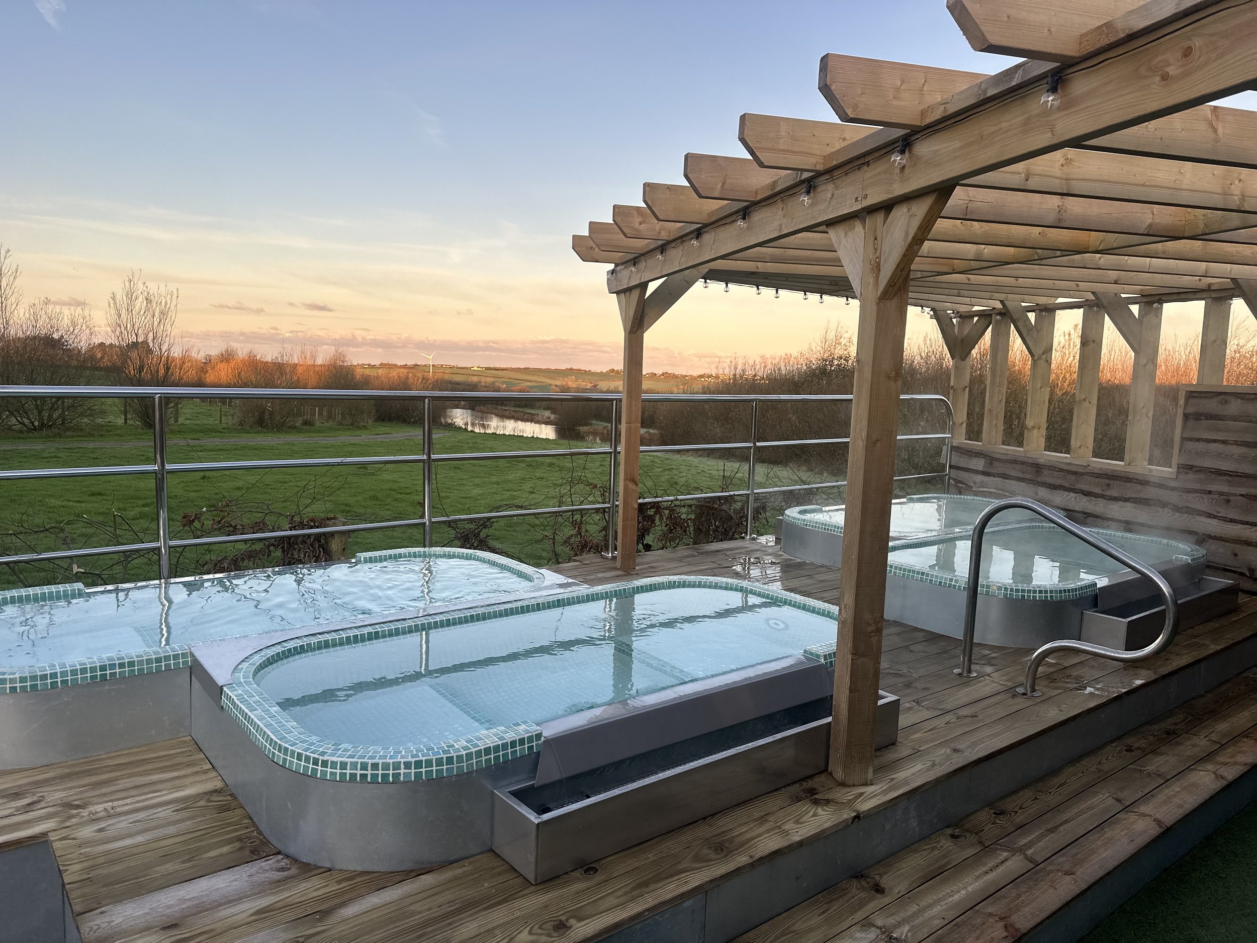 Two outdoor hot tubs on a wooden deck, with a view of a grassy landscape, trees, and a wind turbine in the distance during sunset.
