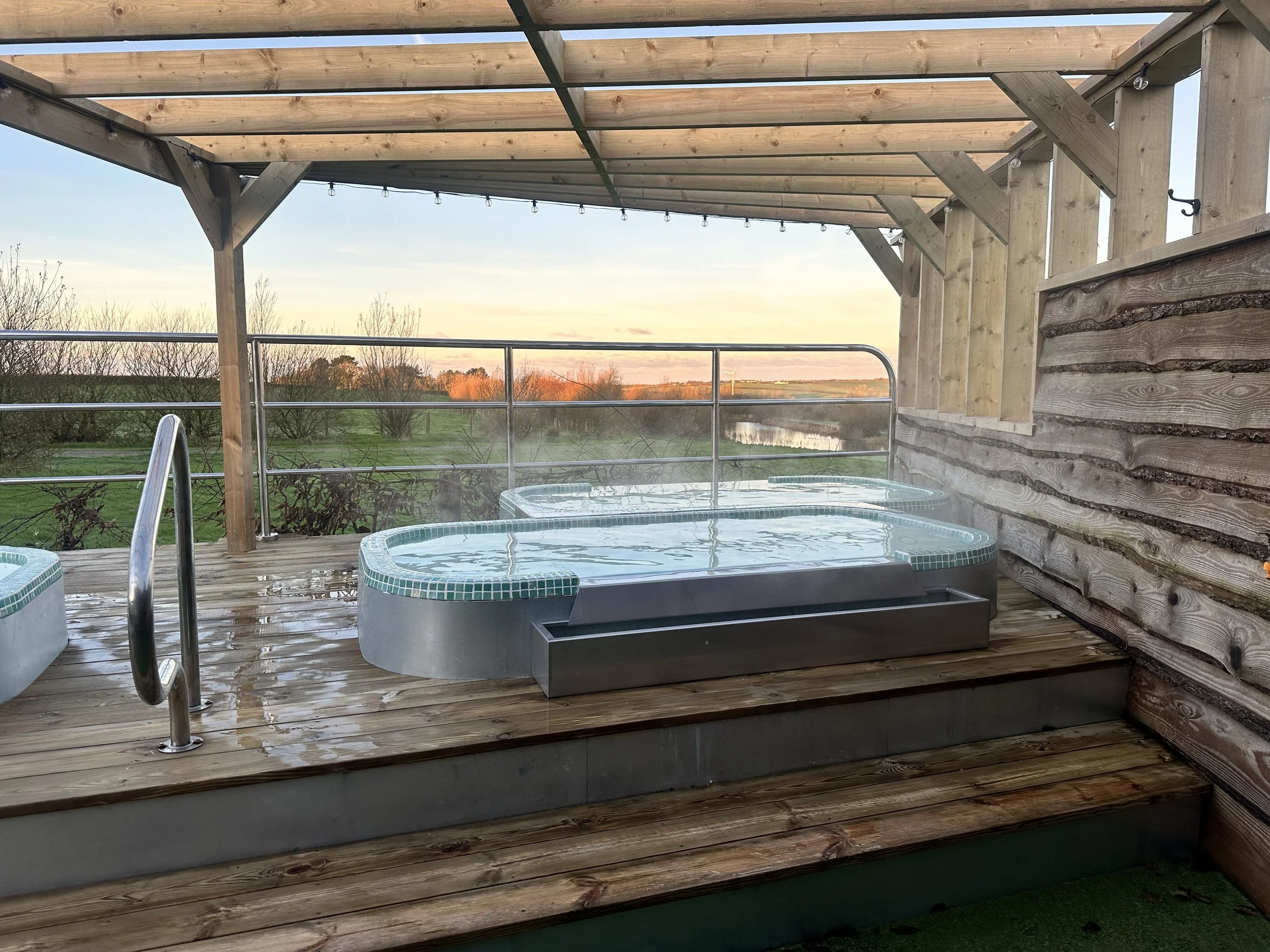 A wooden deck with a hot tub and a partial view of a landscape including trees and a sunset sky.