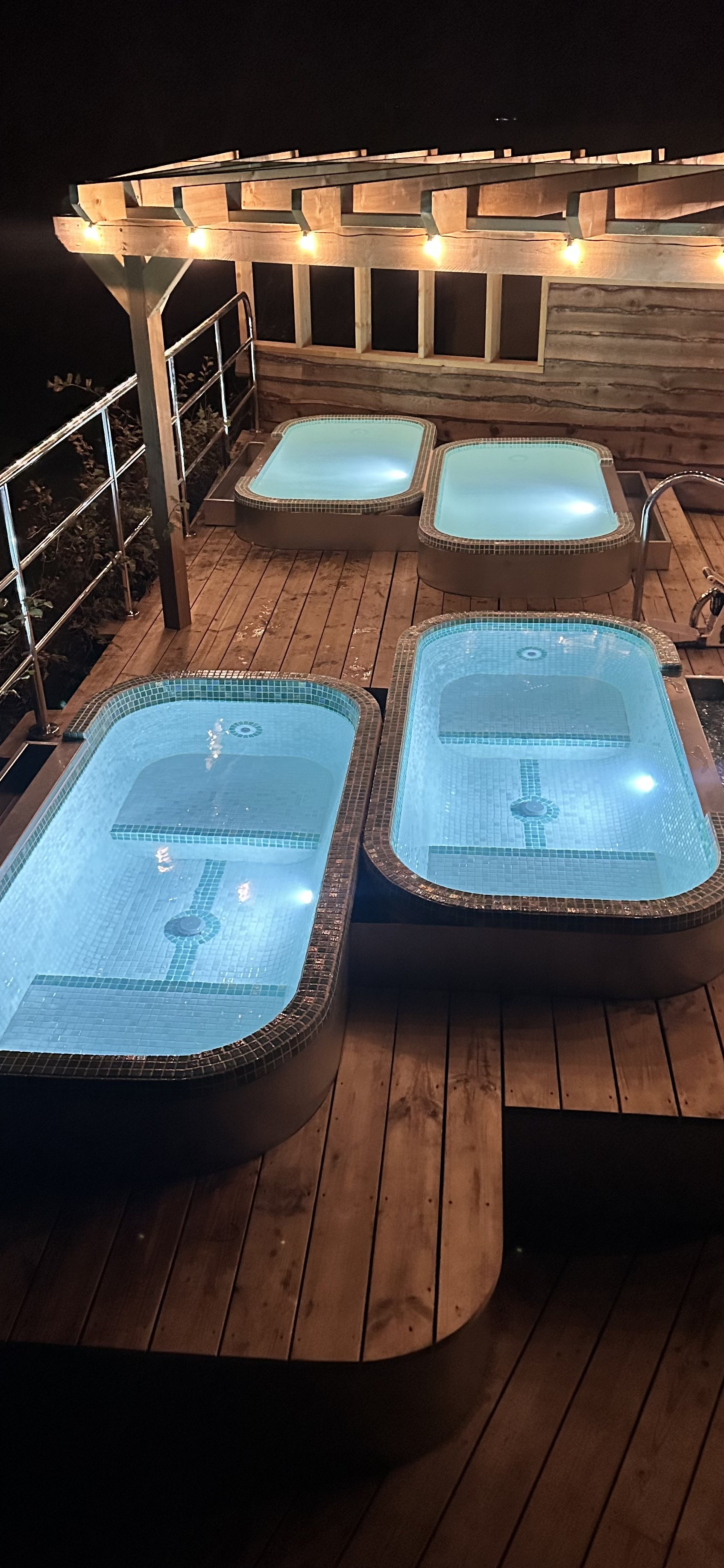Nighttime view of four lit-up hot tubs on a wooden deck with a wooden railing and string lights above.