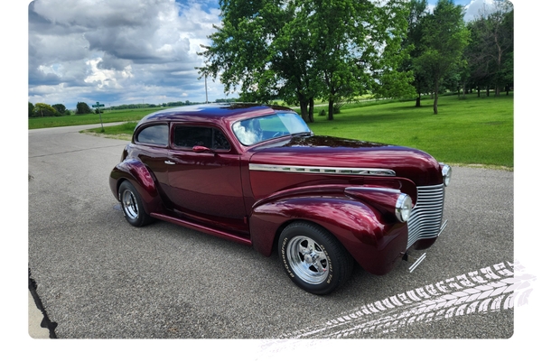 Vintage maroon car parked on a paved road with green trees and grass in the background, cloudy sky overhead.