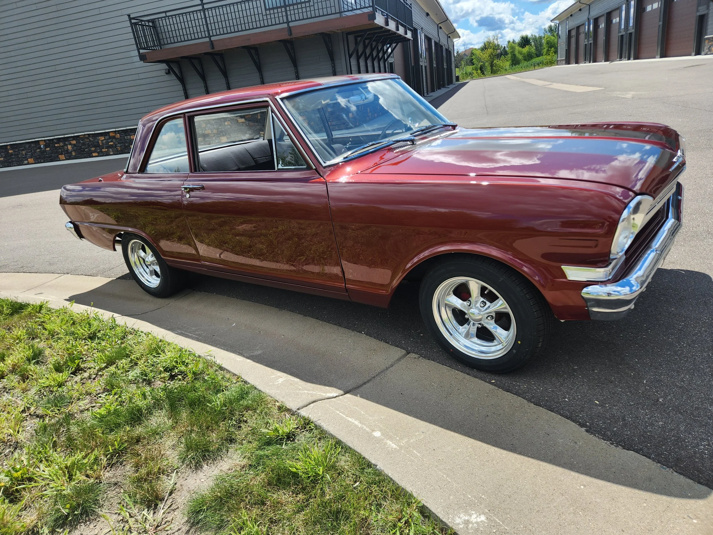 A 1962 Chevy II coupe car with a small block chevy engine and holley carburetor parked beside a sidewalk and grass, with a building in the background, on a sunny day. The car was completed by Truth Dyno and Performance