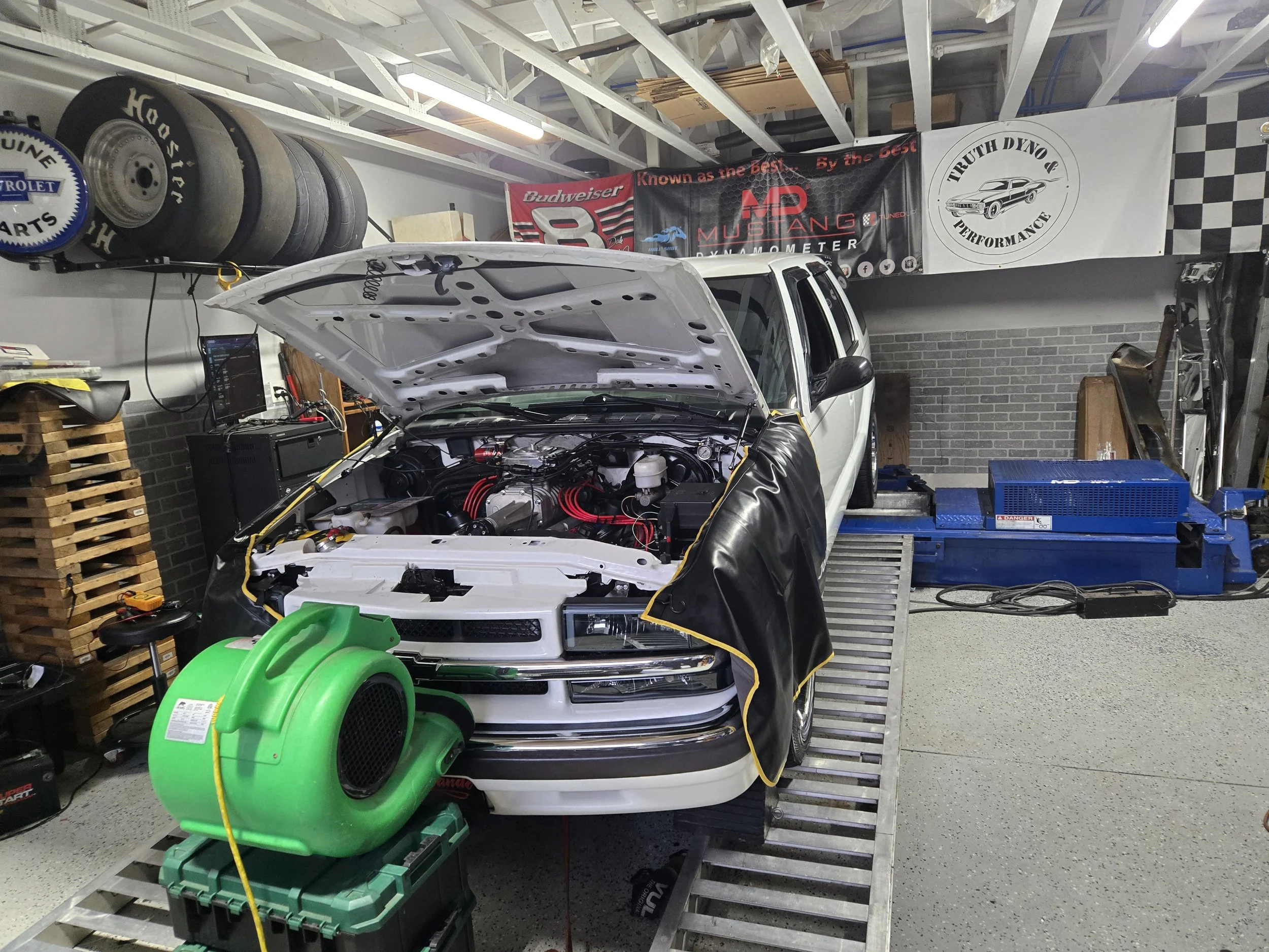 A white pickup truck with its hood open in a garage, showing an engine bay with red spark plug wires and various parts. A green portable air blower is placed in front of the truck. The garage walls have banners for automotive brands and performance parts, with shelves containing tires, tools, and equipment around.