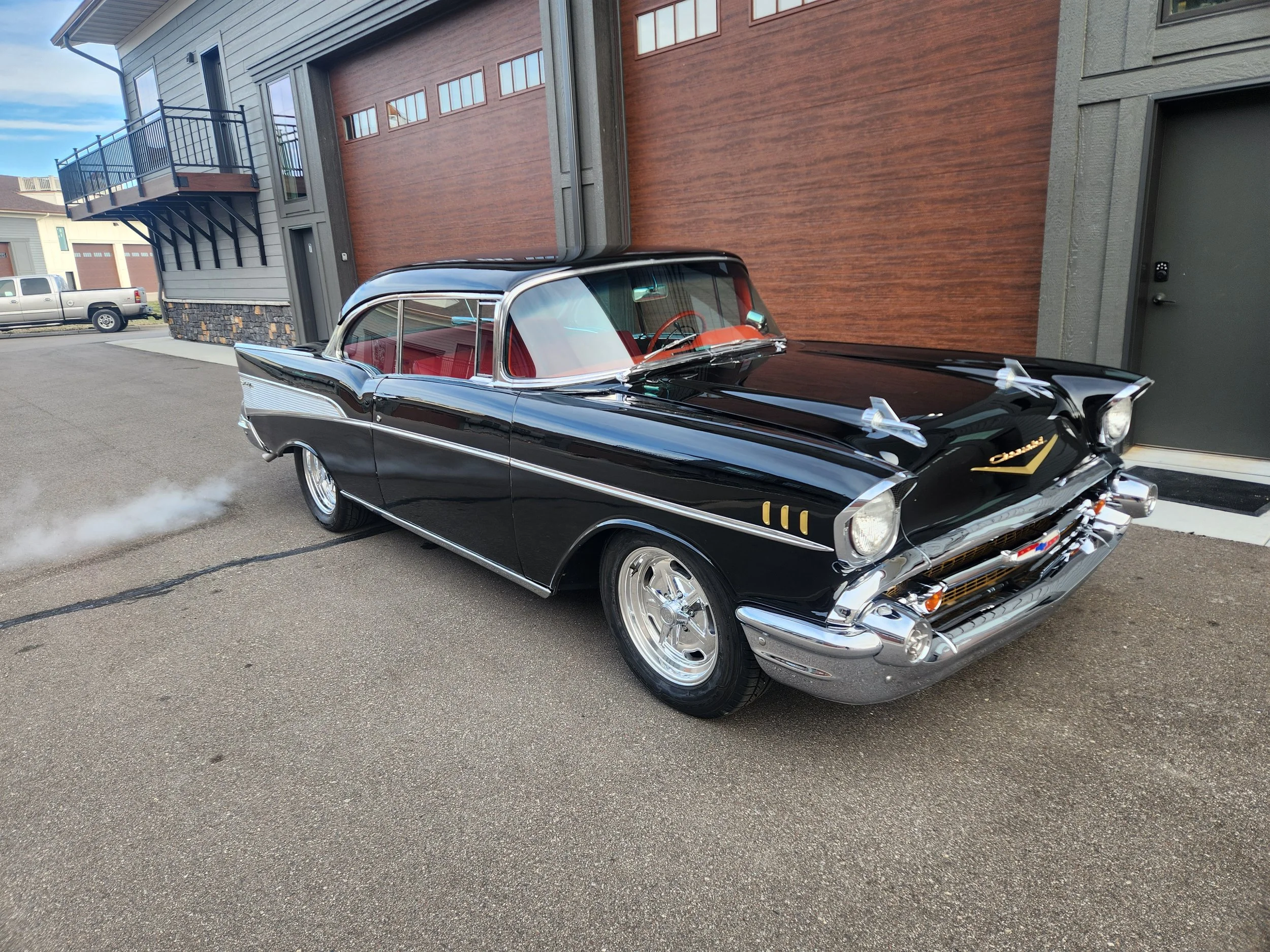 A restored black and white vintage 1957 Chevrolet BelAir car with red interior, gold accents, a 400 cubic inch small block chevy engine from Blueprint Engines, and Holley Carburetor parked outside a building with wooden and gray siding.