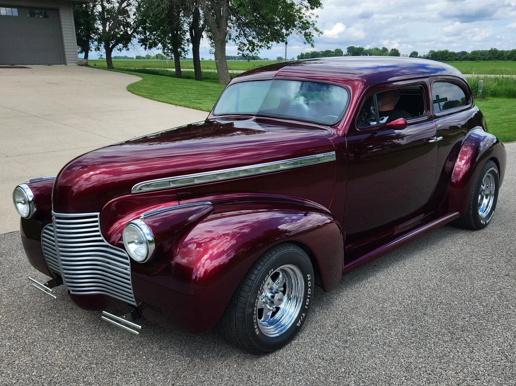 Maroon vintage 1939 Chevy coupe with chopped top, small block chevy engine, and EFI parked on a paved driveway with grassy landscape and trees in the background.
