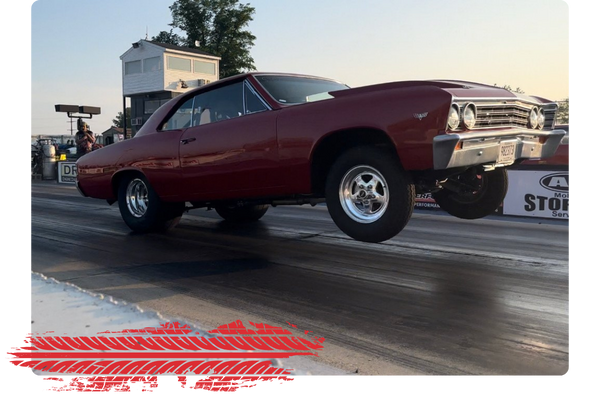 Red vintage mid-1960s muscle car with modified front end, lifting off the ground on a drag racing track.