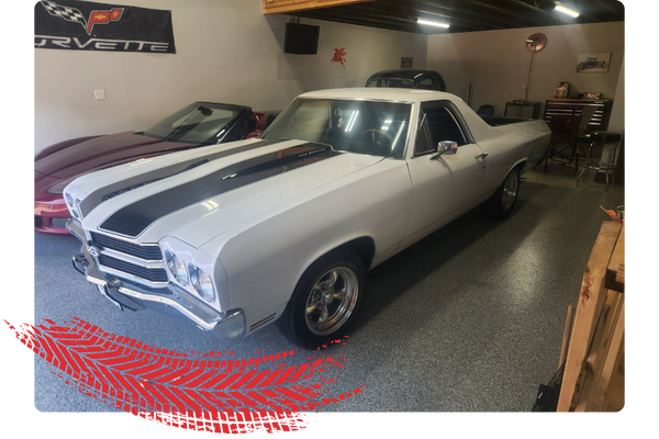 White vintage muscle car with black racing stripes in a garage, alongside other cars, with a Corvette banner on the wall.
